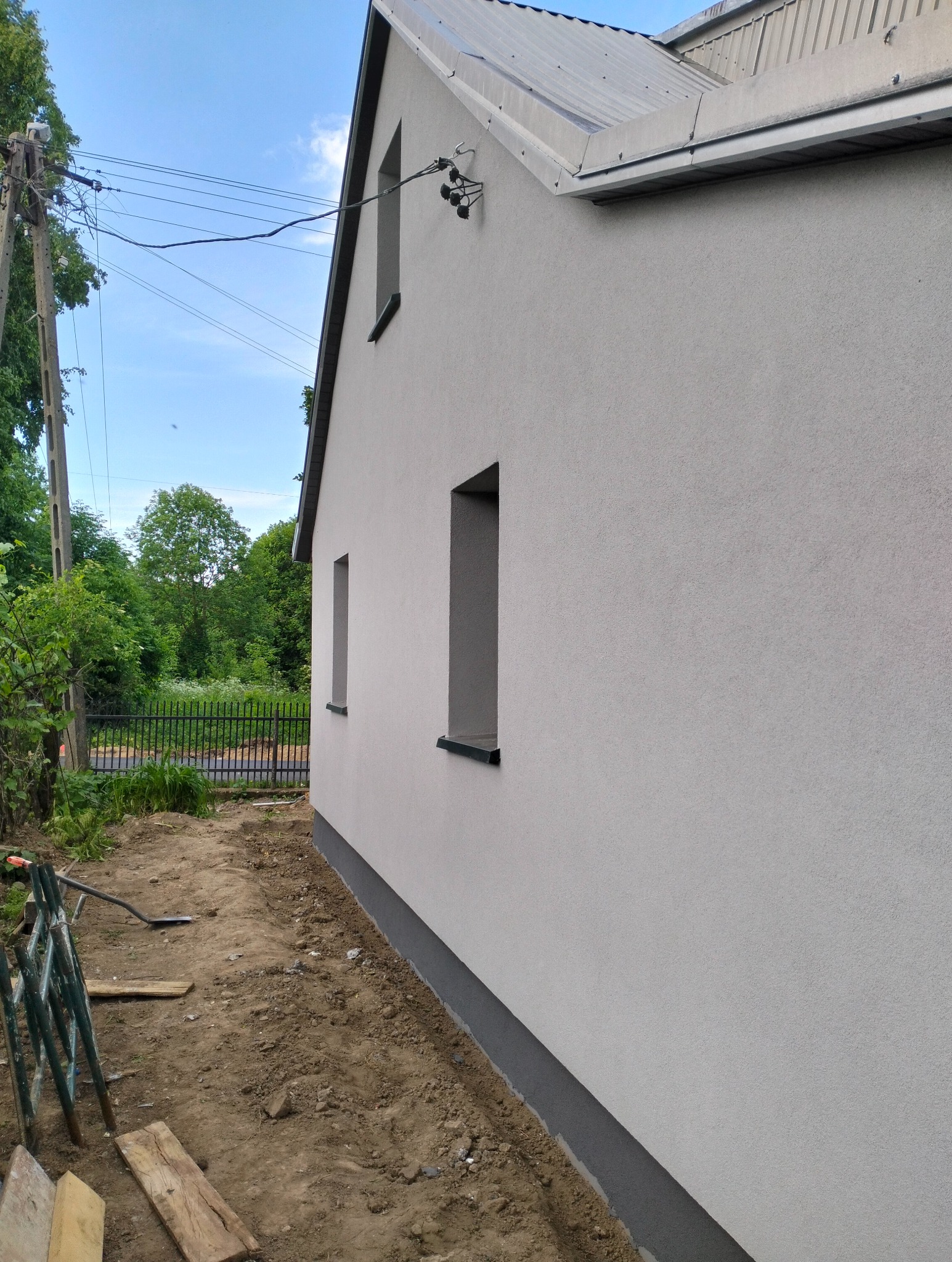 Świeżo otynkowana elewacja domu jednorodzinnego z widocznymi dwoma oknami, szarą podmurówką i fragmentem dachu z blachy trapezowej; na pierwszym planie widoczne rusztowanie i teren wokół domu po...