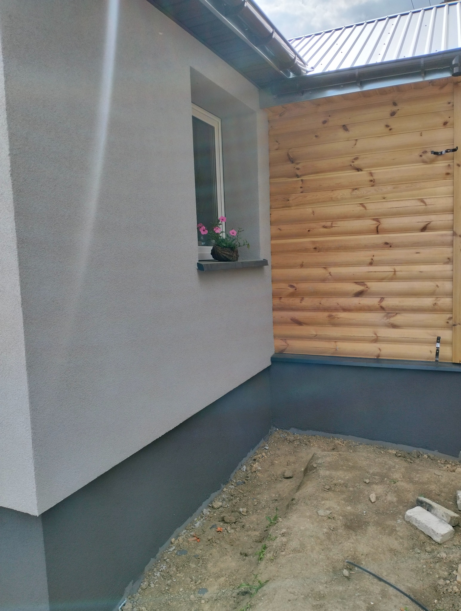 Fragment elewacji domu z oknem, parapetem i doniczką z kwitnącymi różowymi kwiatami, obok drewniana ściana działowa, widoczna szara podmurówka i ziemia.