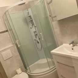 And&Pat Bud - Nowoczesna kabina prysznicowa z hydromasażem i deszczownicą, umywalka z szafką w jasnej łazience wykończonej płytkami imitującymi kamień, widoczny fragment toalety z papierem toaletowym.