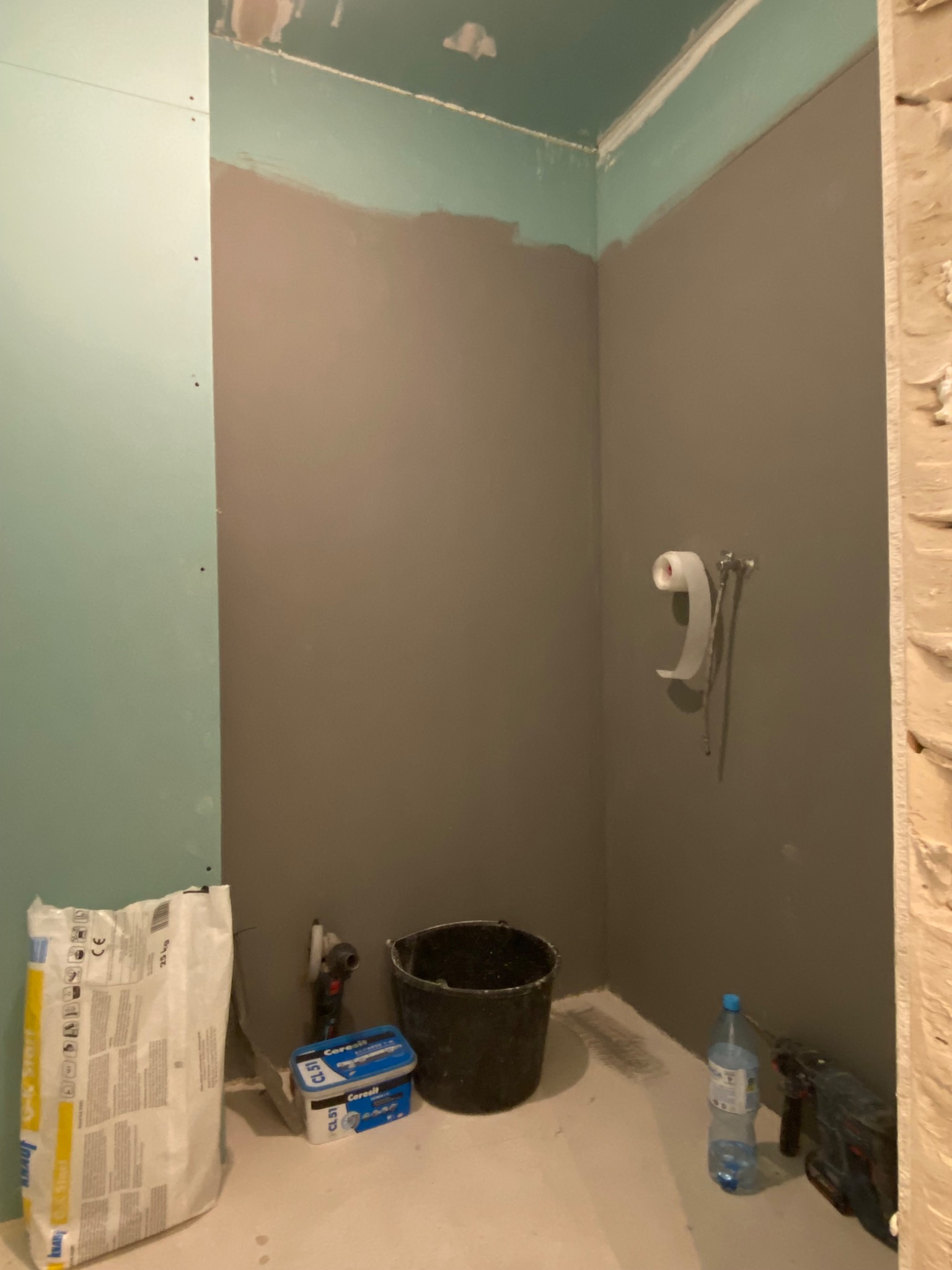 Wnętrze łazienki w trakcie remontu: częściowo położona gładź, widoczny worek z klejem, wiadro, butelka wody oraz narzędzia.