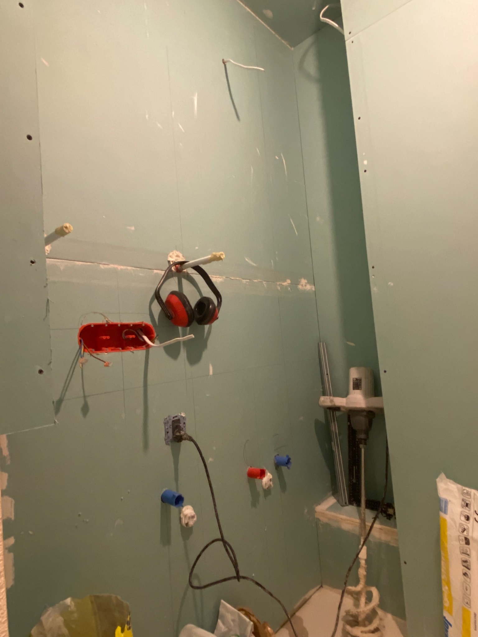 Łazienka w trakcie remontu: ściany pokryte płytami gipsowo-kartonowymi, widoczne puszki elektryczne, słuchawki ochronne na ścianie, mieszadło do zaprawy i worki z zaprawą na podłodze.