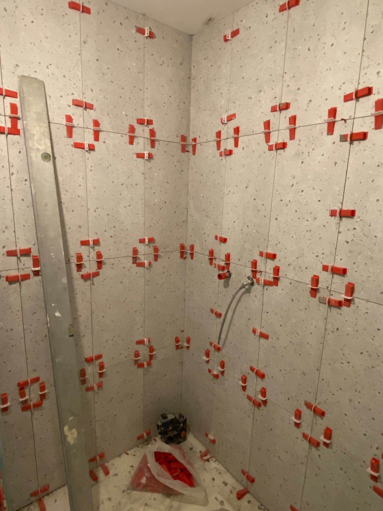 Ściany łazienki w trakcie układania płytek z użyciem systemu poziomowania płytek (klipsy i kliny), widoczna poziomica i narzędzia glazurnicze.