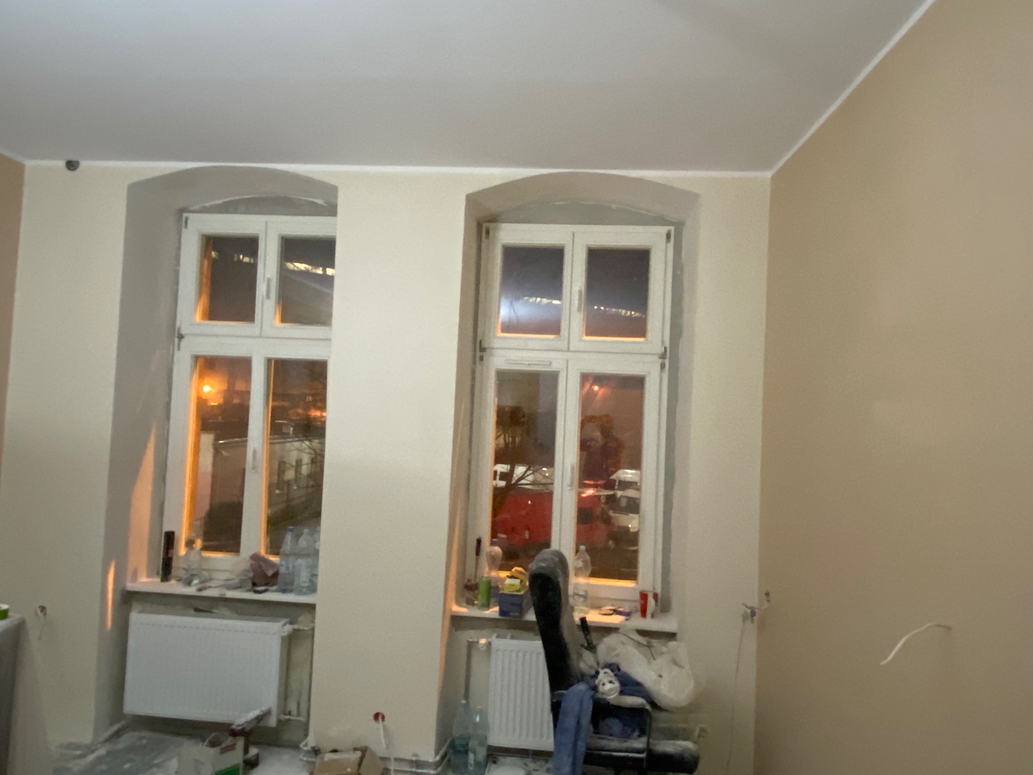 Pokój w trakcie malowania ścian na beżowo, widoczne dwa okna z zaokrąglonymi górnymi krawędziami, grzejniki pod oknami, butelki z wodą i inne przedmioty na parapecie, krzesło obok okna.