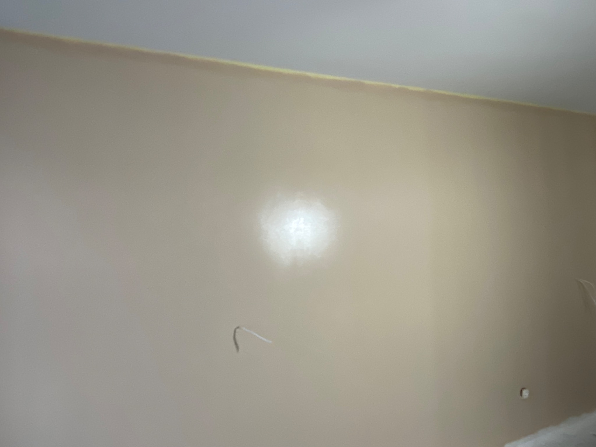 Świeżo pomalowana ściana w odcieniach beżu, z widoczną taśmą malarską przy suficie i wystającymi przewodami elektrycznymi, gotowa do dalszych prac wykończeniowych.