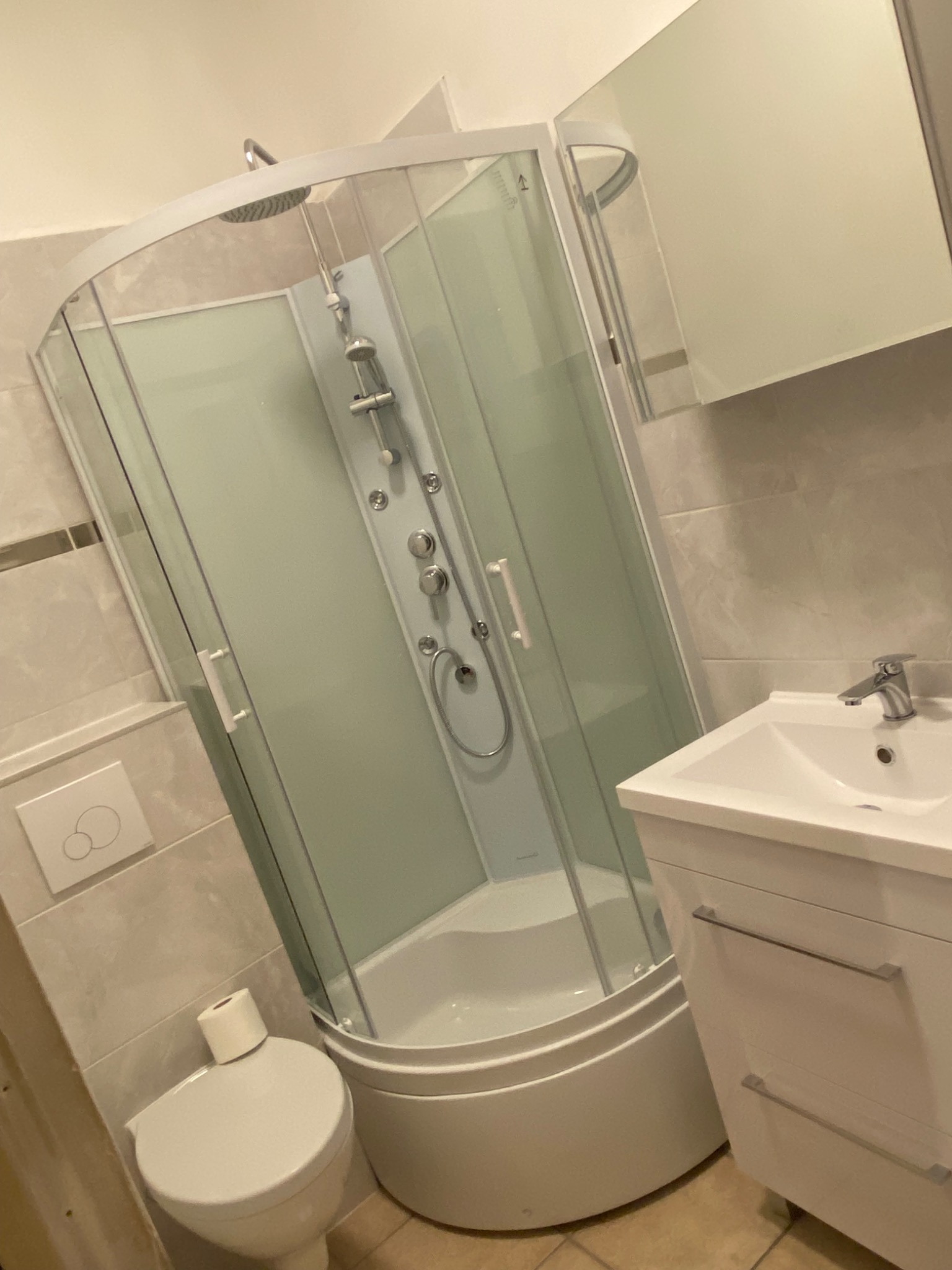 Nowoczesna kabina prysznicowa z hydromasażem i deszczownicą, umywalka z szafką w jasnej łazience wykończonej płytkami imitującymi kamień, widoczny fragment toalety z papierem toaletowym.