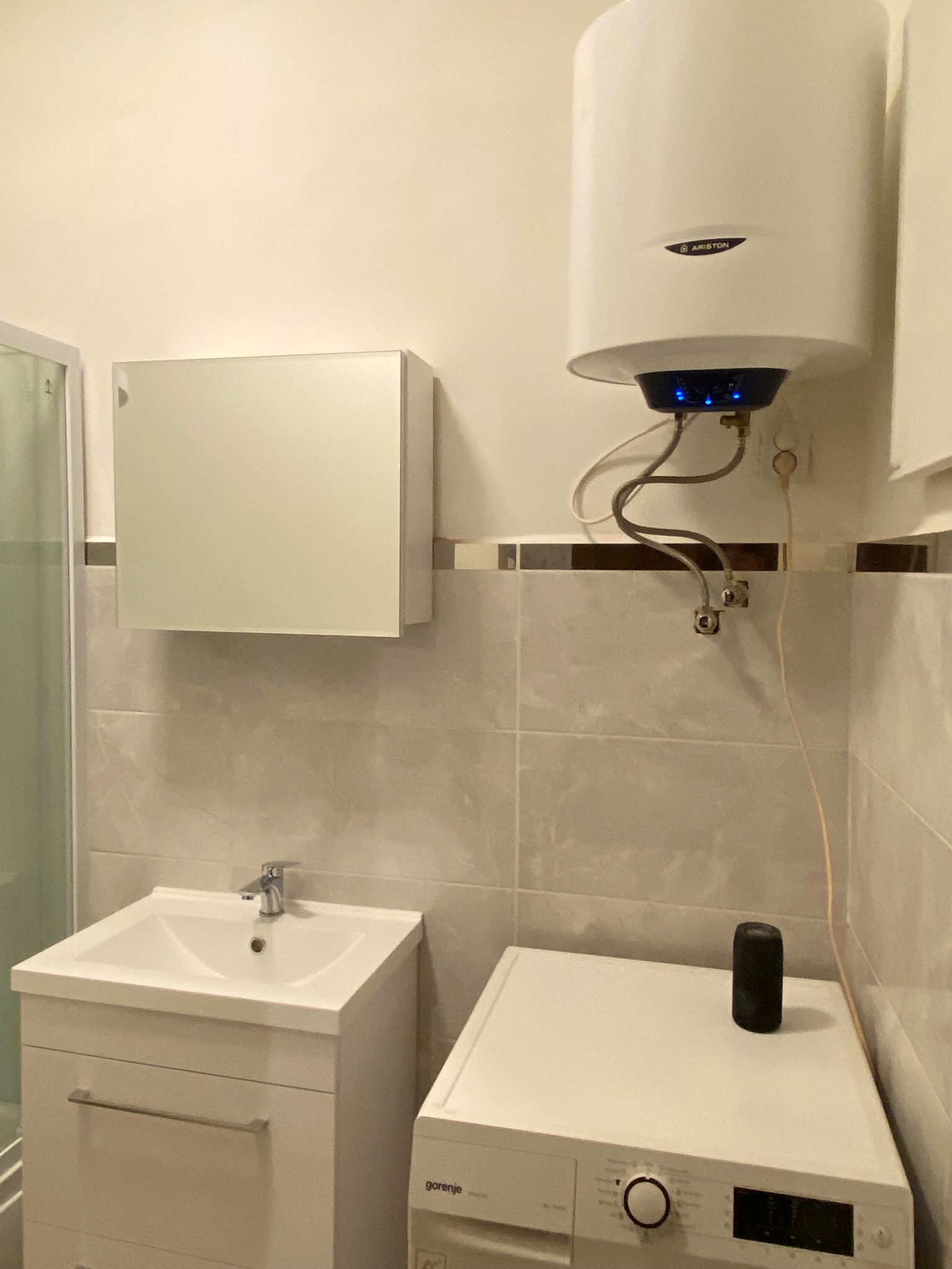 Biała łazienka z umywalką w szafce, pralką Gorenje i wiszącym bojlerem Ariston z podświetleniem LED, płytki w odcieniach beżu.
