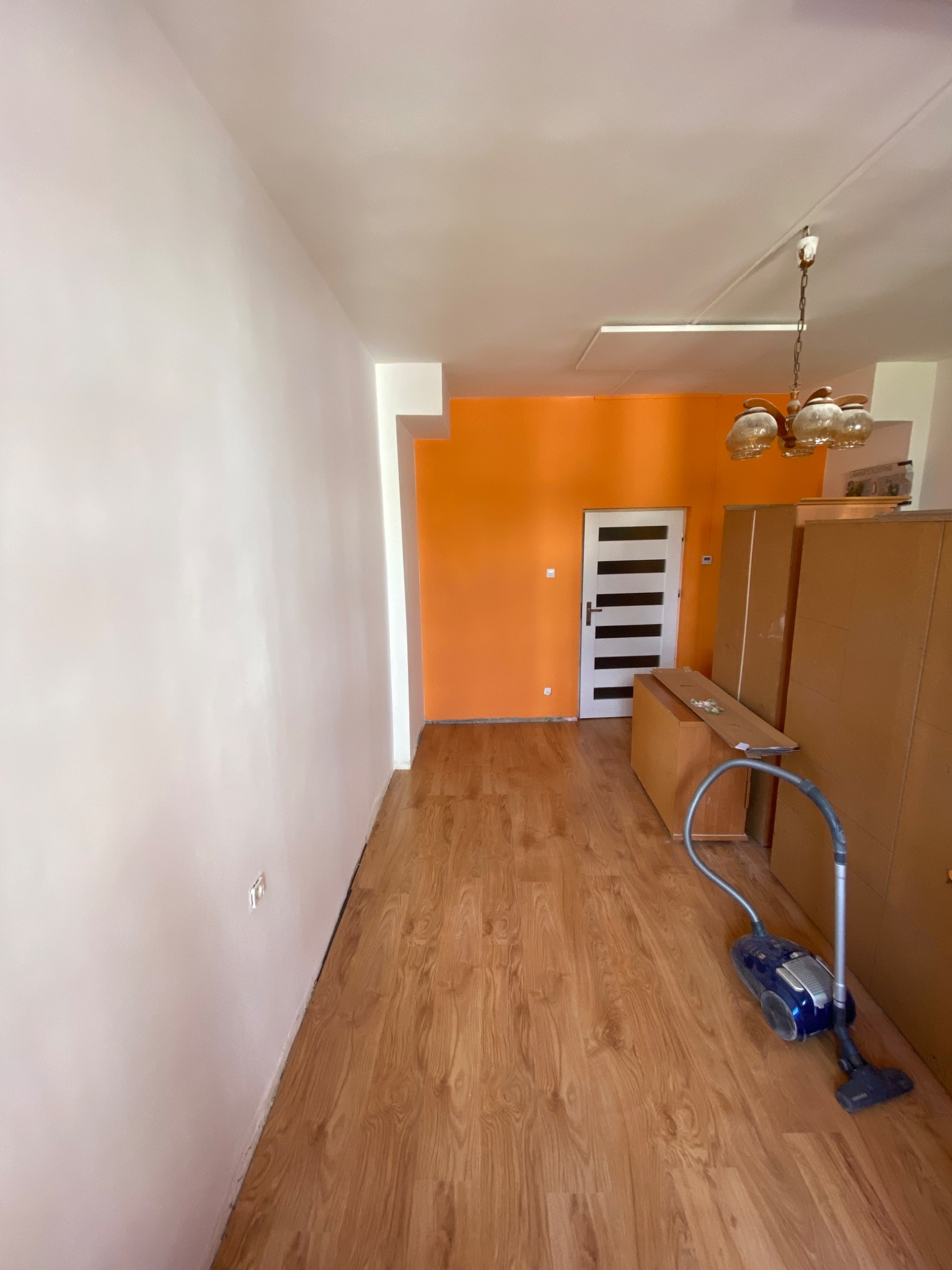 Pokój po remoncie z odkurzaczem na podłodze, pomarańczowa ściana i białe drzwi w tle, drewniana podłoga, widoczne szafy i lampa sufitowa.
