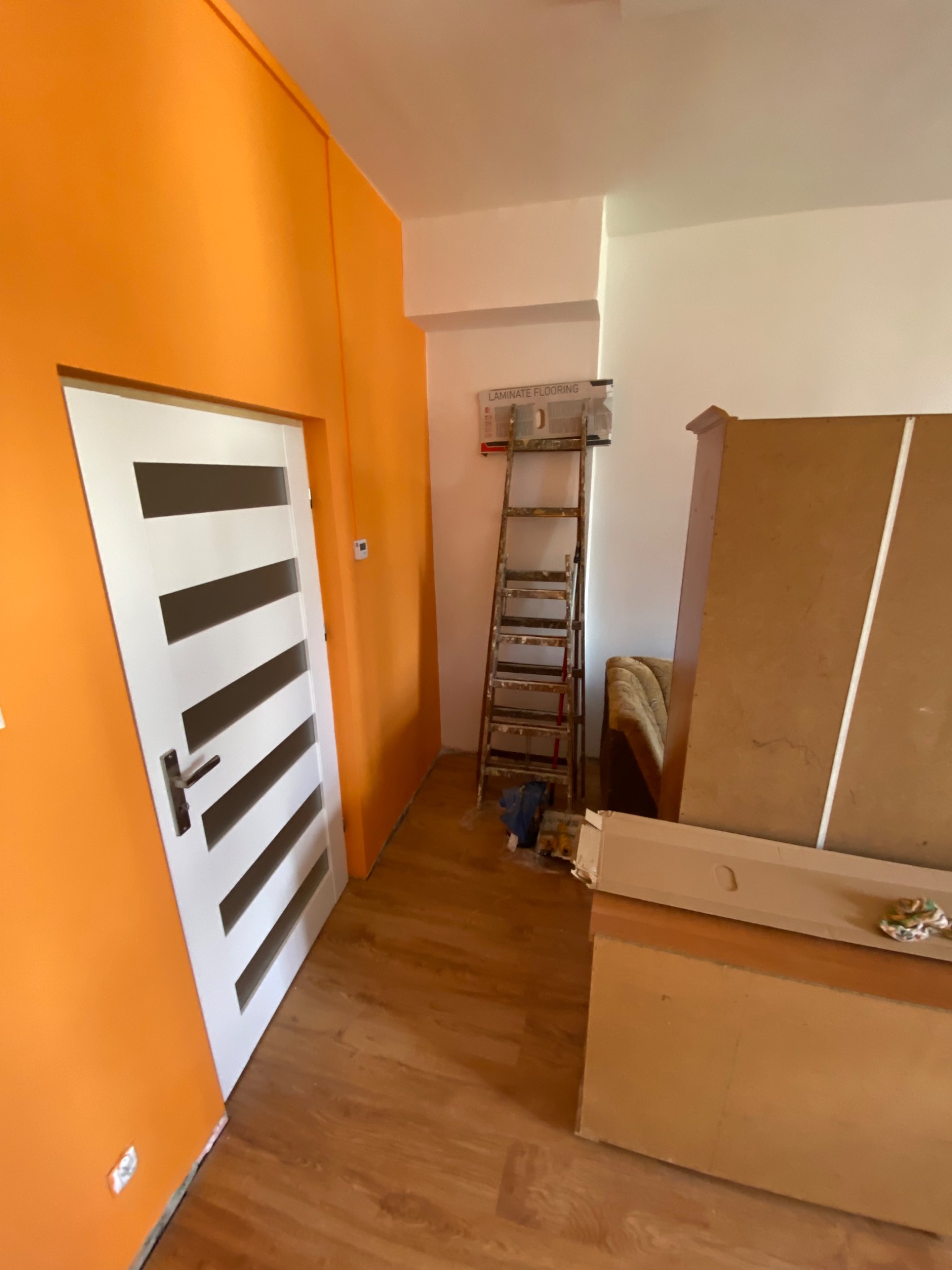 Pomarańczowa ściana po malowaniu, białe drzwi z poziomymi, ciemnymi paskami, drabina malarska i rozpakowane panele podłogowe w trakcie prac wykończeniowych.