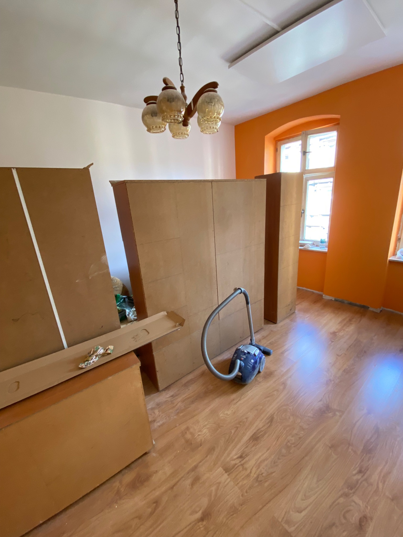 Pokój w trakcie wykończenia: rozpakowane elementy mebli, odkurzacz na parkiecie, pomarańczowa ściana z oknem i wiszący żyrandol.