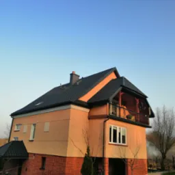 Dwupiętrowy dom z ciemną dachówką, balkonem i elewacją w odcieniach beżu i czerwieni, sfotografowany w słoneczny dzień.