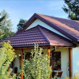 Dom jednorodzinny z brązowym dachem z blachodachówki, widoczny komin ze stali nierdzewnej i rynny w kolorze dachu, otoczony zielenią drzew i krzewów.