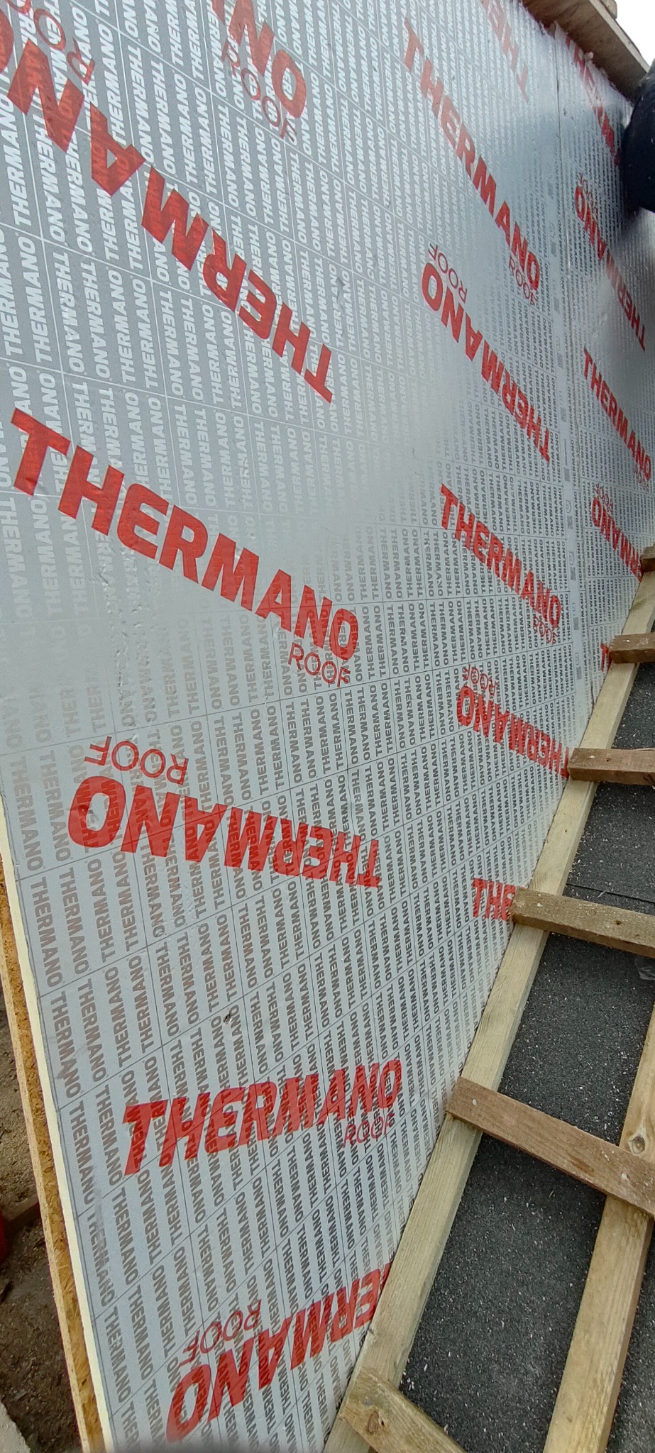 Widok z góry na fragment dachu w trakcie montażu, pokrytego membraną dachową z wyraźnym nadrukiem 'THERMANO ROOF', z widocznymi łatami dachowymi i fragmentem czarnego pokrycia.