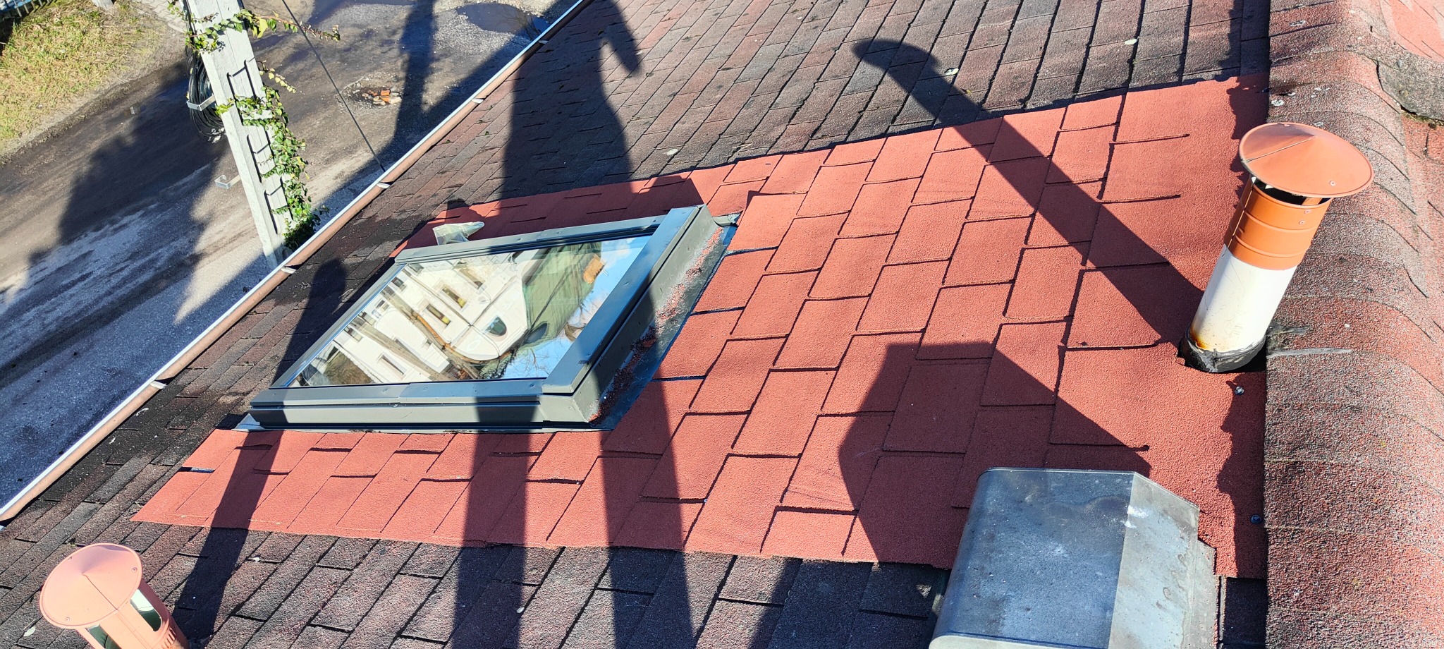 Fragment dachu pokryty czerwonymi gontami bitumicznymi, z zamontowanym oknem dachowym z szarą ramą i kominem wentylacyjnym z pomarańczowym daszkiem, cień drzewa na powierzchni.