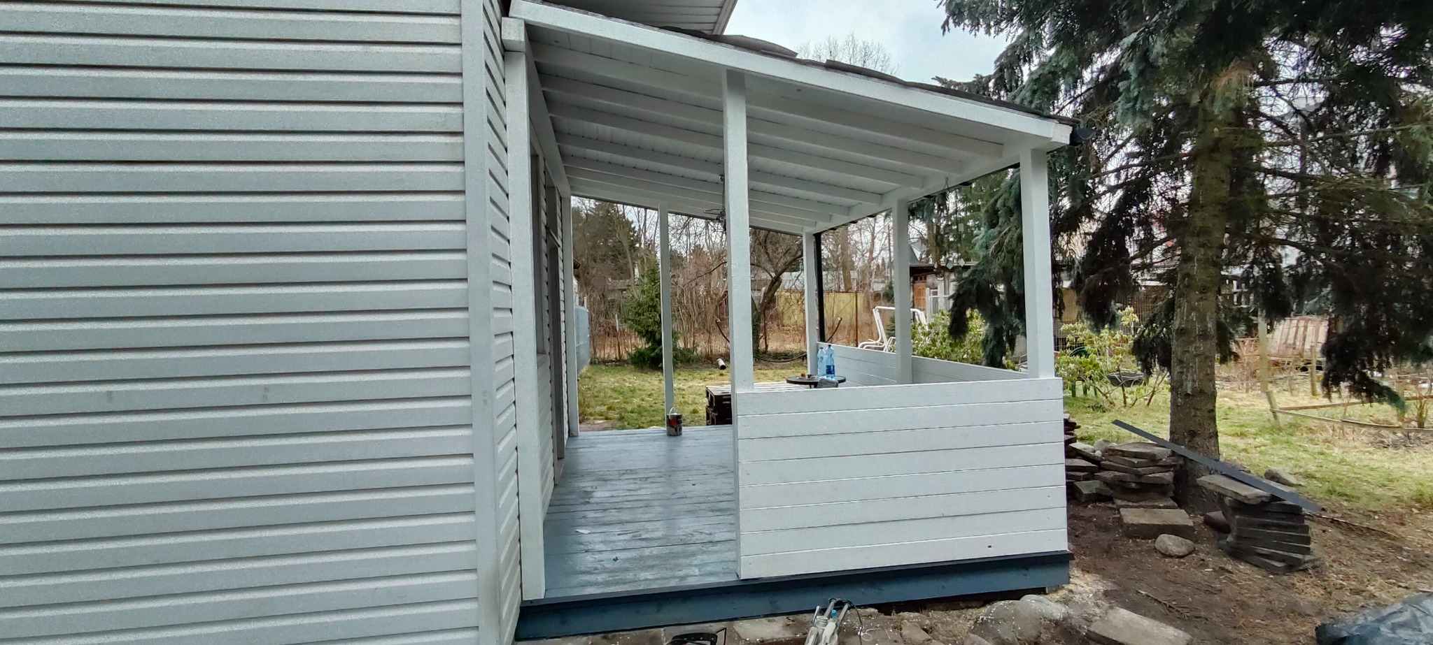 Boczny widok na zadaszony ganek z białymi słupami i drewnianą balustradą, przylegający do domu z szarą elewacją sidingową; na pierwszym planie stos kamiennych płyt.