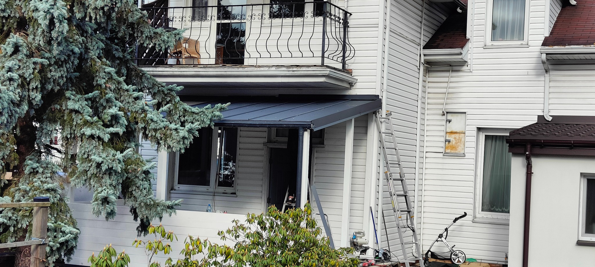 Dom z sidingu z nową, grafitową obróbką blacharską nad wejściem, drabina oparta o ścianę, widoczny fragment rynny i iglaste drzewo z przodu.