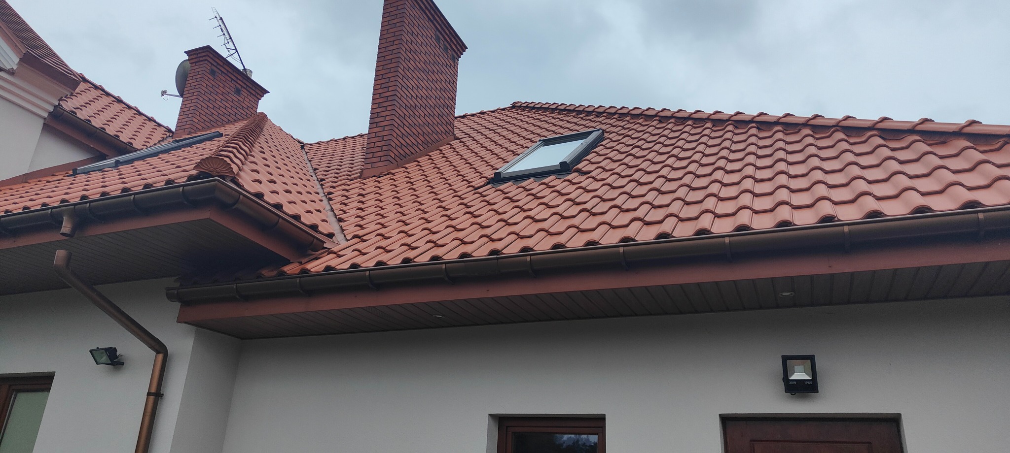 Czerwony dach ceramiczny z oknem dachowym i brązowymi rynnami na tle pochmurnego nieba, widoczny komin z cegły i antena.