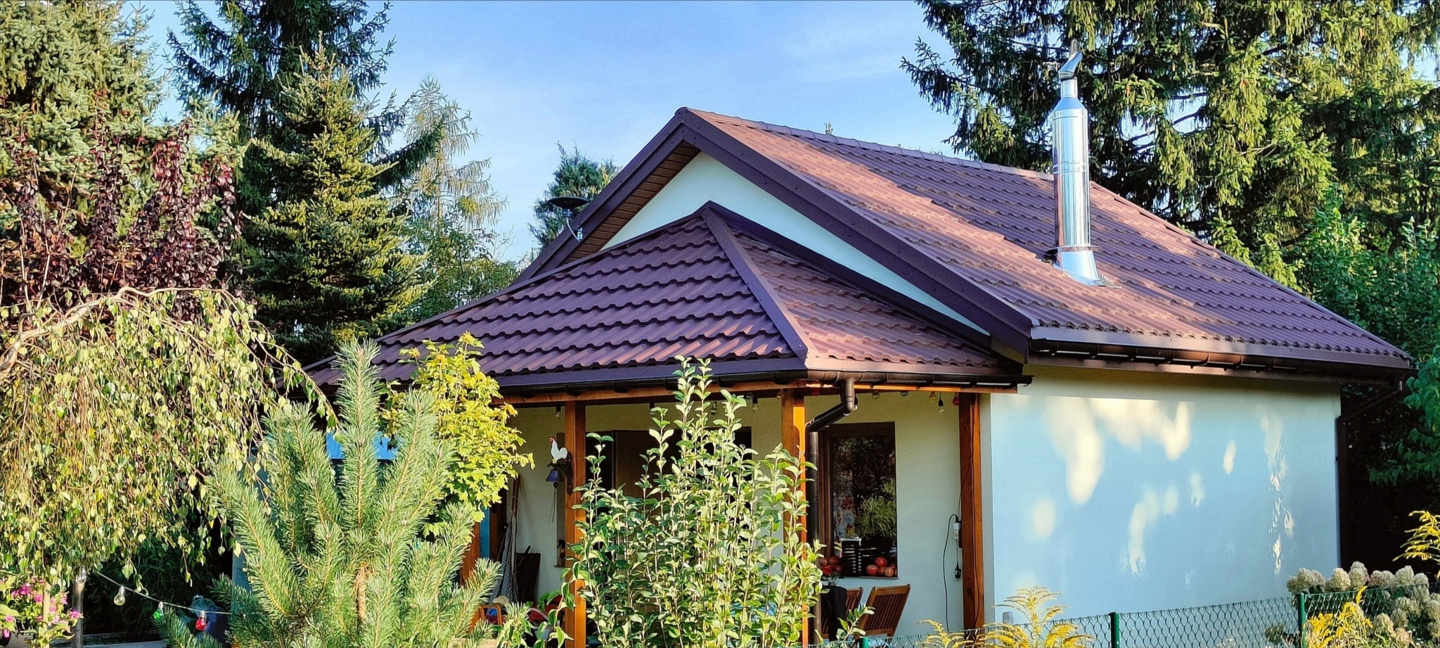 Dom jednorodzinny z brązowym dachem z blachodachówki, widoczny komin ze stali nierdzewnej i rynny w kolorze dachu, otoczony zielenią drzew i krzewów.