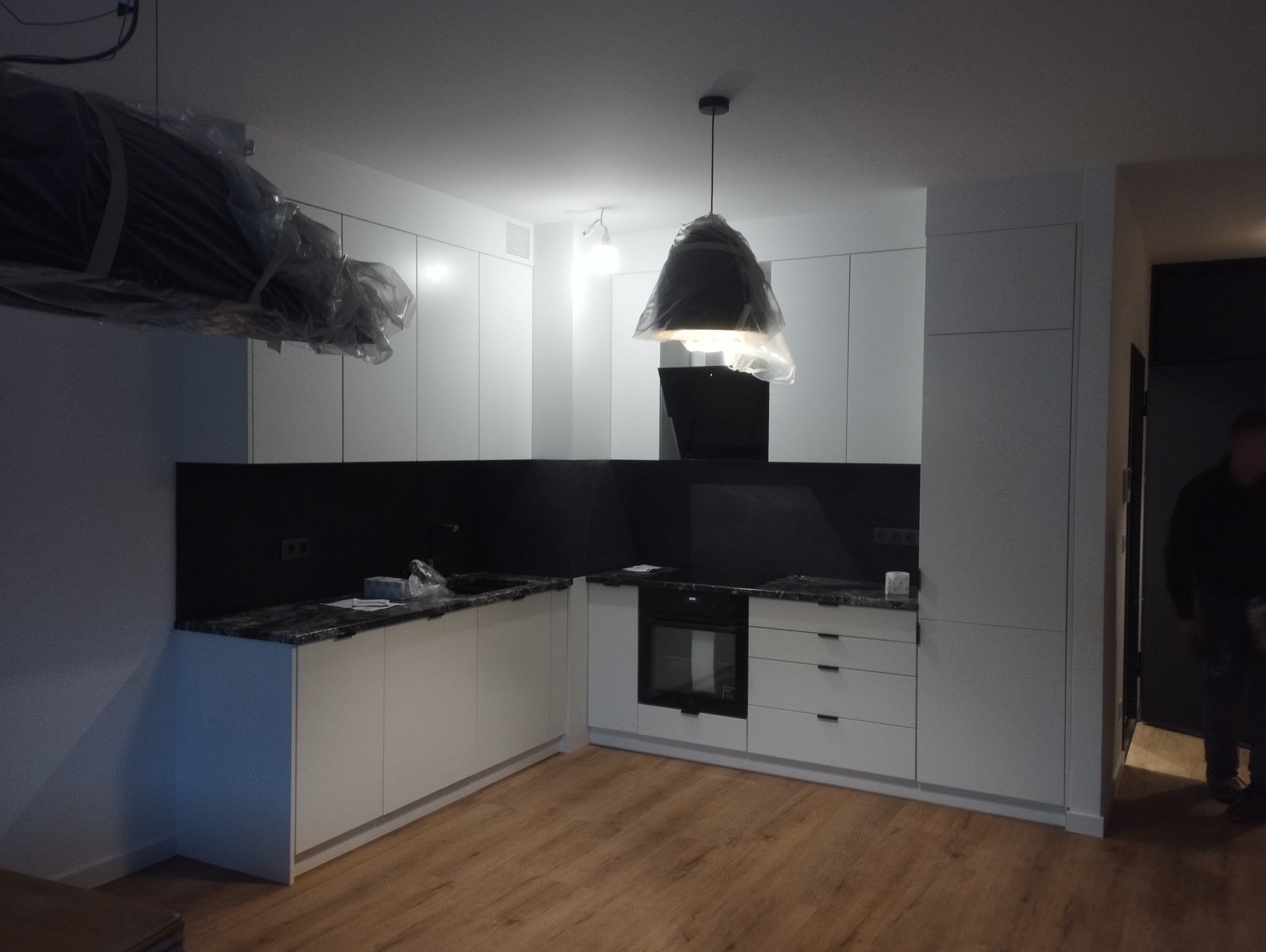 Nowoczesna kuchnia z białymi szafkami i czarnym blatem, jeszcze w folii ochronnej, z widocznym oświetleniem i drewnianą podłogą, w trakcie montażu lub tuż po nim.