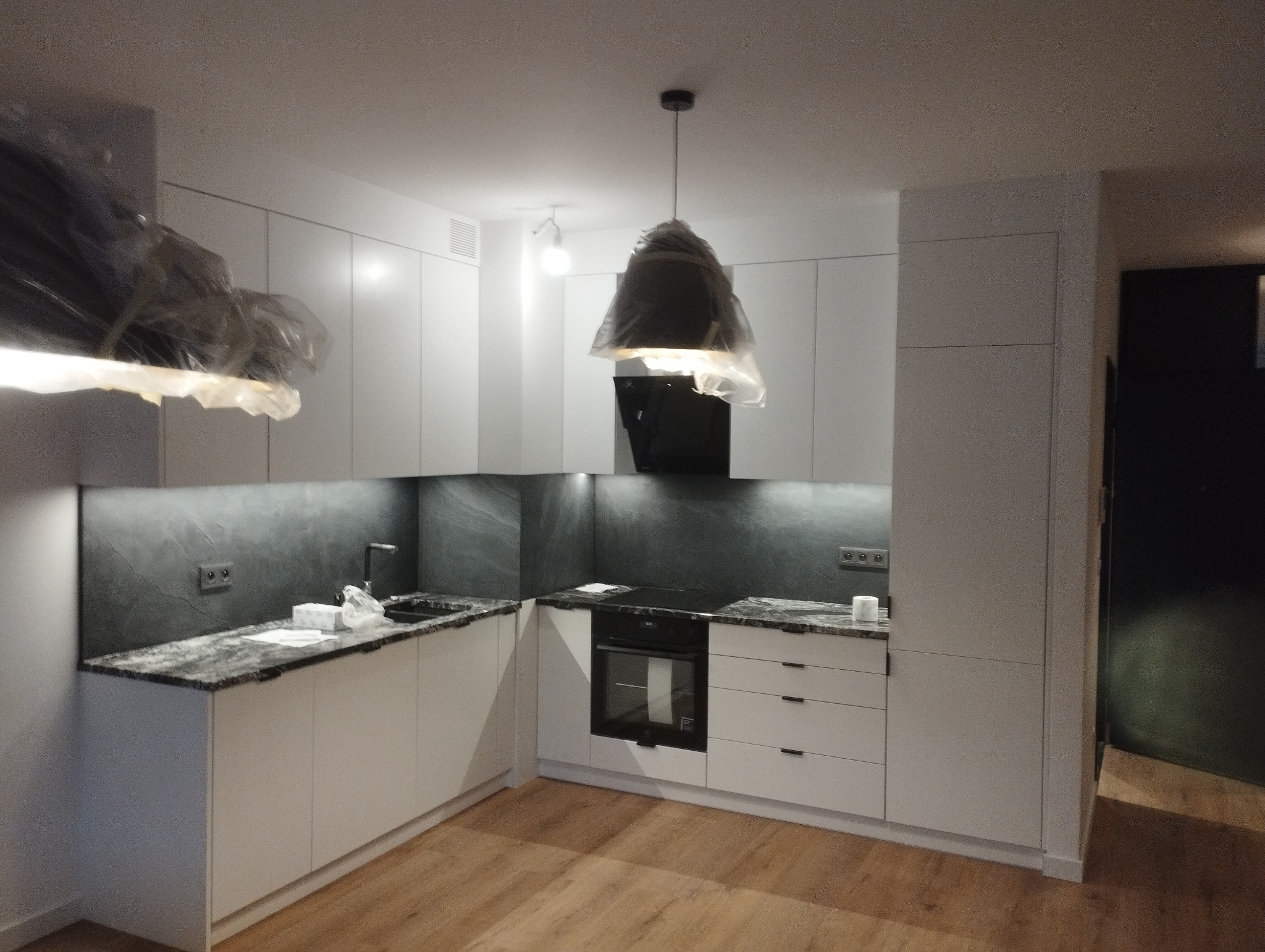 Nowoczesna kuchnia z białymi szafkami, blatem imitującym kamień, czarnym okapem i lampami wiszącymi zabezpieczonymi folią ochronną, na podłodze panele.