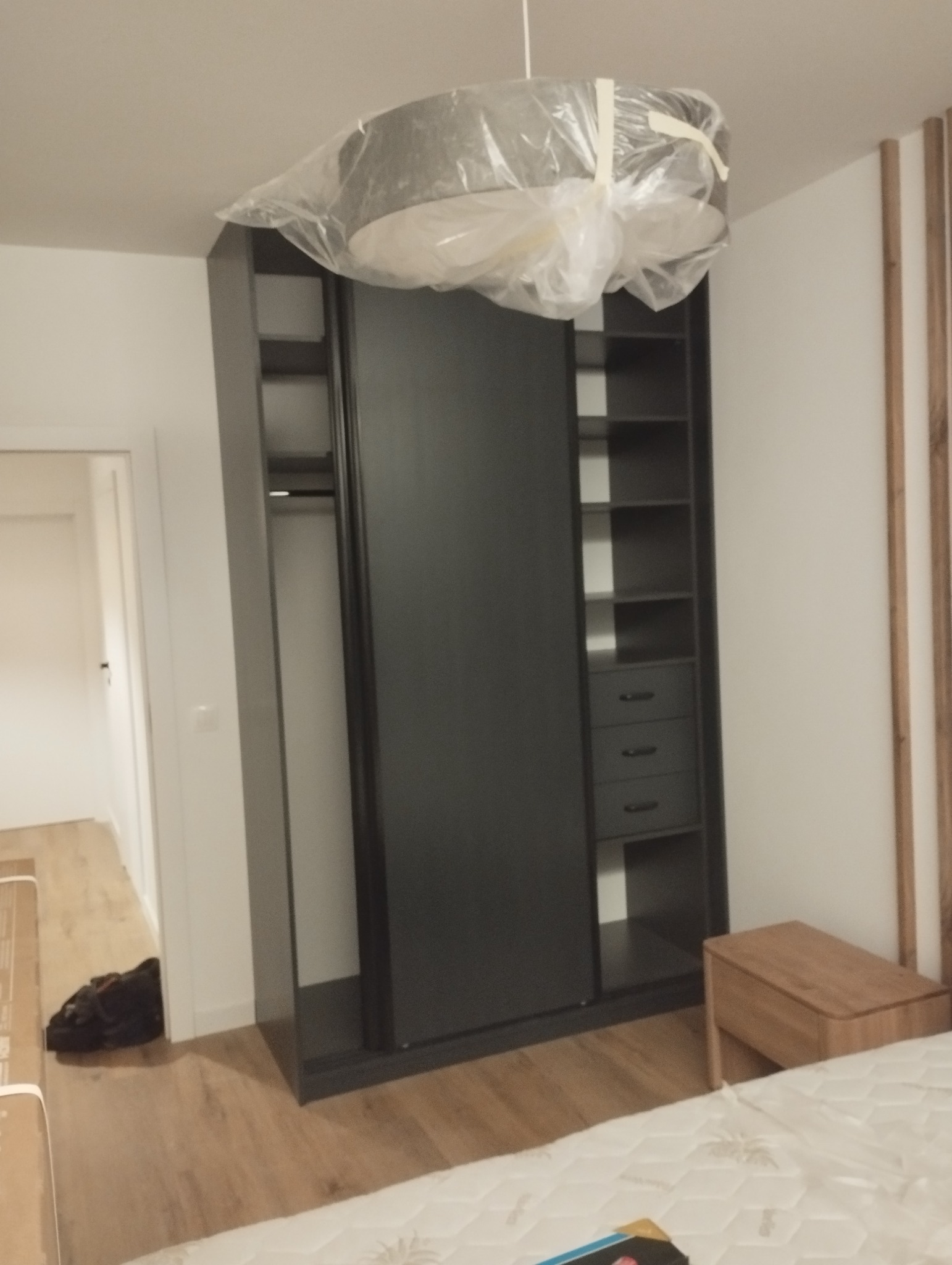 Czarna szafa przesuwna z półkami i szufladami, widok z boku, lampa wisząca zabezpieczona folią, podłoga z paneli, fragment łóżka i szafki nocnej