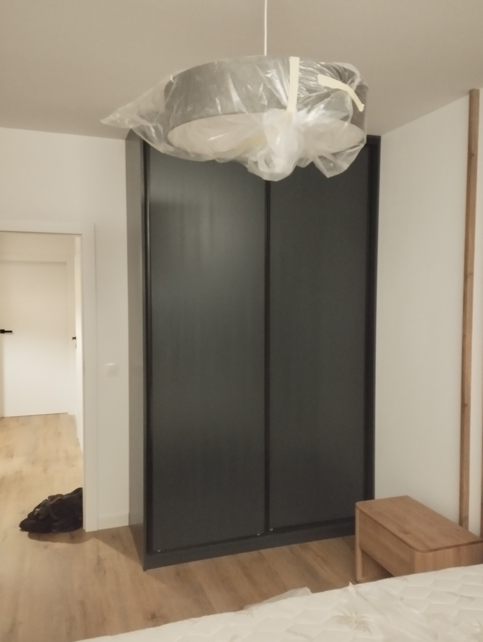 Nowoczesna szafa przesuwna z ciemnymi drzwiami w sypialni, lampa w folii ochronnej, widok na otwarte drzwi do innego pomieszczenia.