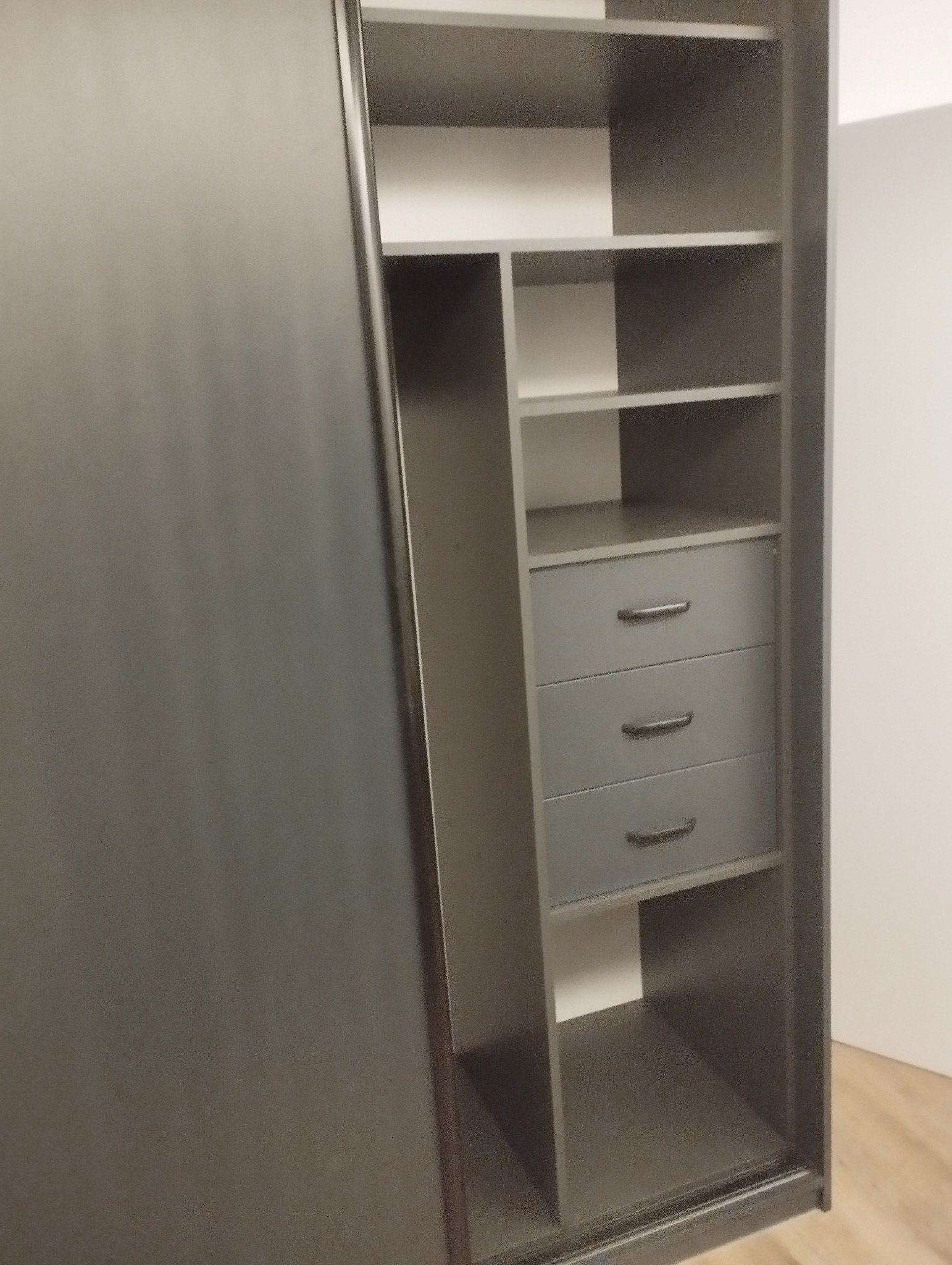 Wnętrze szafy wnękowej z przesuwnymi drzwiami w kolorze grafitowym, widoczne półki i trzy szuflady z ciemnymi uchwytami, minimalistyczny design.