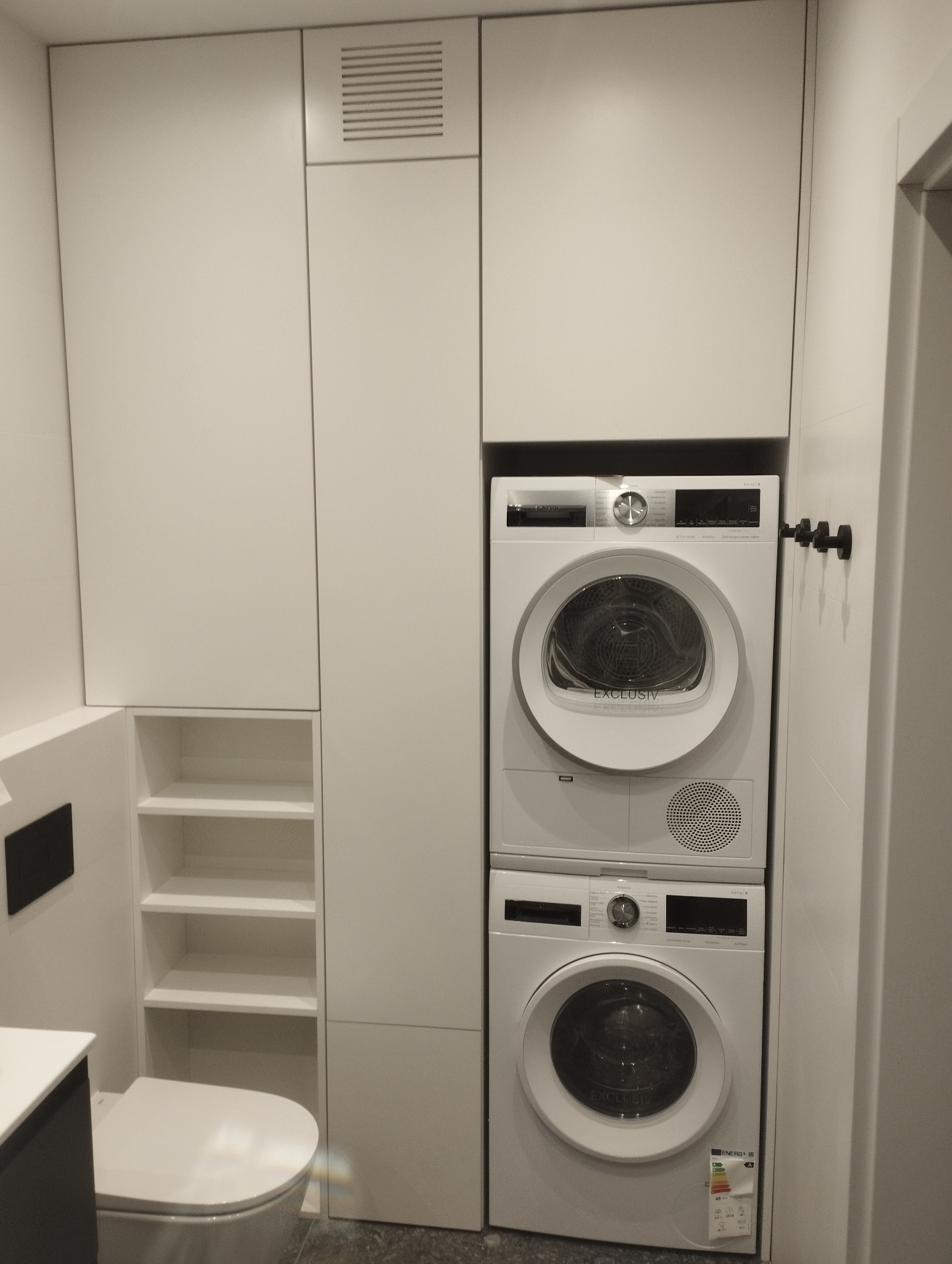 Biała łazienka z zabudową meblową na wymiar, w której umieszczono pralkę i suszarkę w kolumnie. Po lewej stronie toaleta i półki.