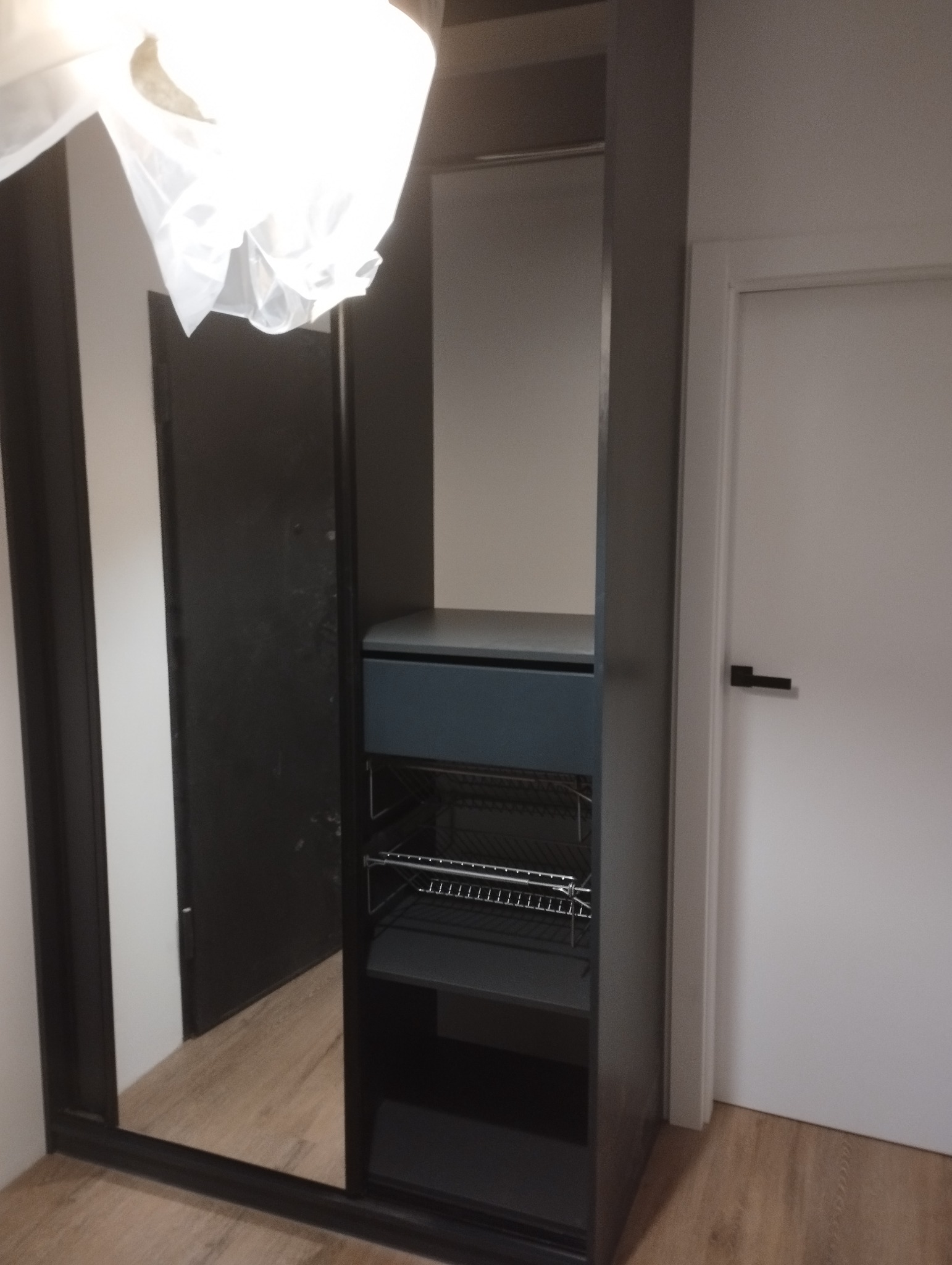 Ciemnoszara szafa na wymiar z szufladą i metalowymi półkami, widoczna w lustrzanym odbiciu, z drzwiami w tle.