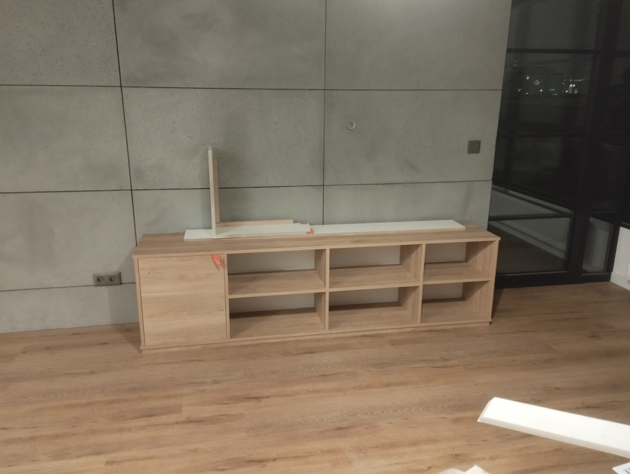 Niedokończona szafka RTV z jasnego drewna na podłodze z paneli, z fragmentami białych elementów na blacie, na tle szarej ściany z betonowych płyt i okna.