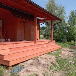 RT-Styl - Nowy, drewniany taras z zadaszeniem przy domu, pomalowany na ciepły, pomarańczowy kolor. Widoczne schody wejściowe i elementy konstrukcyjne. W tle zieleń drzew i błękitne niebo.