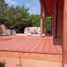 RT-Styl - Ciepły odcień drewna na tarasie z meblami ogrodowymi i relaksującą osobą w tle, otoczony zielenią. Widoczne elementy konstrukcyjne z drewna.