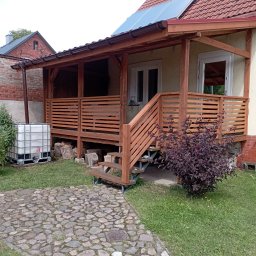 RT-Styl - Zadaszony taras z drewnianą balustradą i schodami, widok z zewnątrz. Obok zbiornik na wodę i ścieżka z kamienia polnego. Na dachu panele słoneczne.