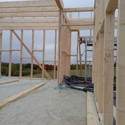 RT-Styl - Wnętrze domu w trakcie budowy z drewnianą konstrukcją szkieletową, widoczne elementy konstrukcyjne i drabina. W tle jesienny krajobraz za oknem.