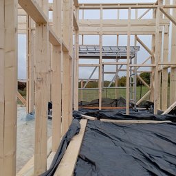 RT-Styl - Surowy szkielet domu w budowie, z widocznymi drewnianymi belkami i rusztowaniem w tle. Czarna folia ochronna na podłożu. Widok z bliska na konstrukcję.