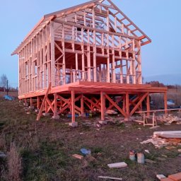 RT-Styl - Szkielet drewniany domu w trakcie budowy, widoczny na tle nieba o zmierzchu. Konstrukcja osadzona na betonowych fundamentach, z widocznymi elementami drewnianymi.