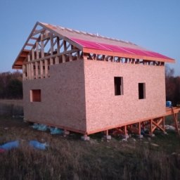 RT-Styl - Dom szkieletowy w trakcie budowy: widoczne ściany z płyt OSB, konstrukcja dachu z różową membraną, fundamenty z bloczków betonowych. Ujęcie z zewnątrz, w tle łąka i drzewa.