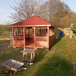 RT-Styl - Drewniana altana z czerwonym dachem w ogrodzie, w trakcie montażu. Widoczny stół roboczy, taczka z drewnem i drabina. W tle drzewa i ogrodzenie.