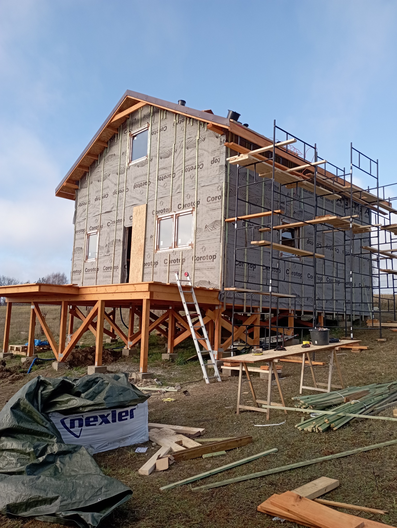 Dwu-kondygnacyjny dom szkieletowy w trakcie budowy, z widocznym ociepleniem ścian, rusztowaniami i drewnianym tarasem na podwyższeniu. Widoczne materiały budowlane.