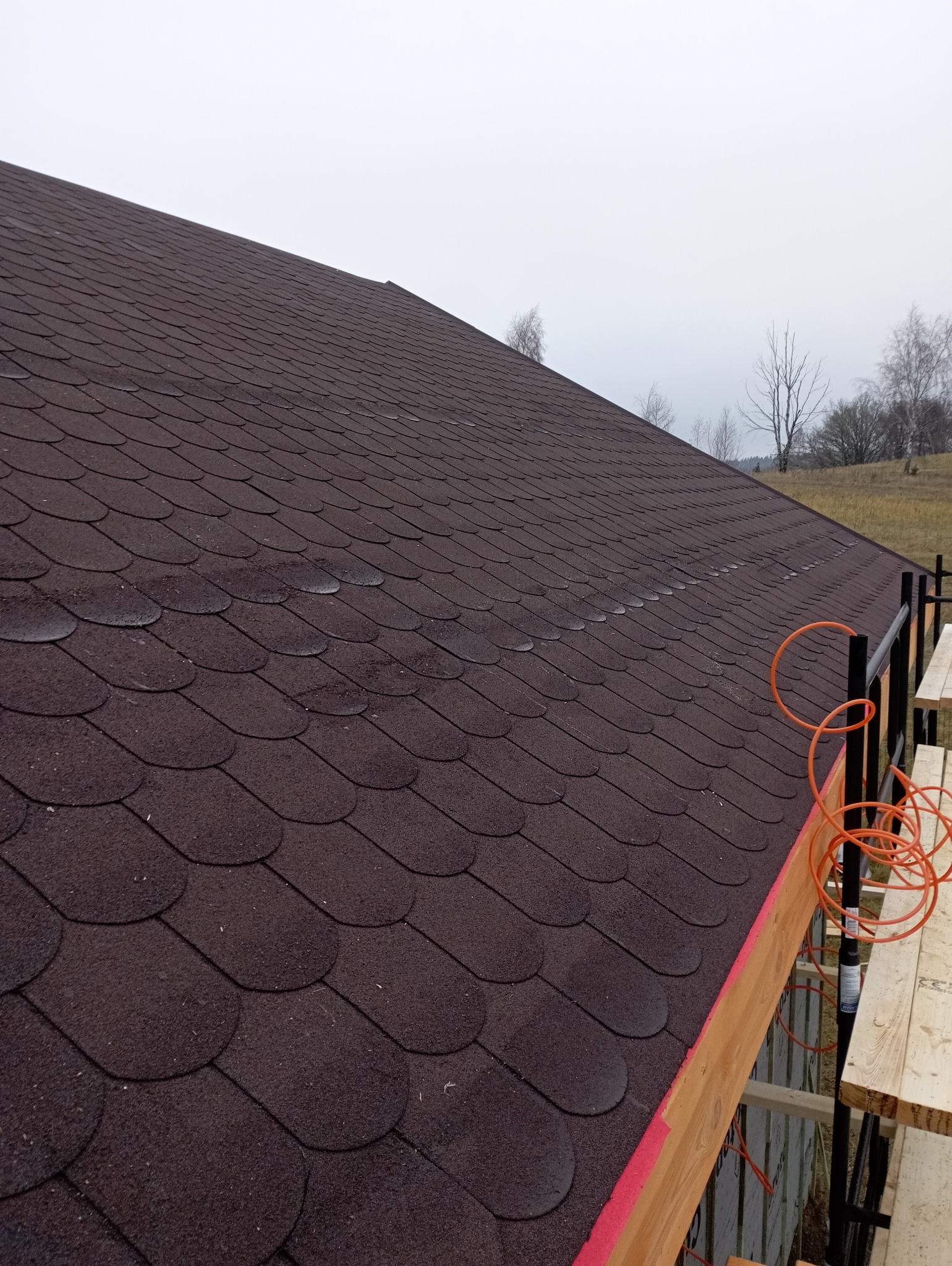 Brązowy dach pokryty gontem bitumicznym, widoczny ze rusztowania. W tle drzewa i pochmurne niebo. Ujęcie z boku, ukazujące strukturę i kolor dachu.
