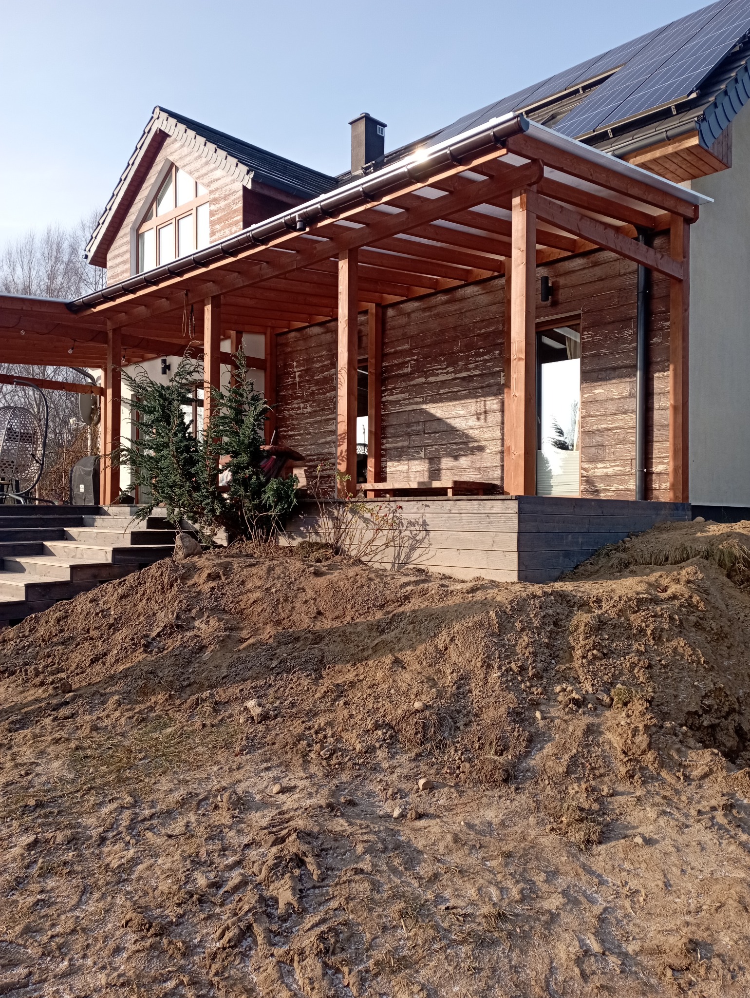 Drewniana pergola z zadaszeniem przy domu, widok z dołu na konstrukcję i fragment elewacji. Uwagę zwraca ziemia przed tarasem, sugerująca prace wykończeniowe.