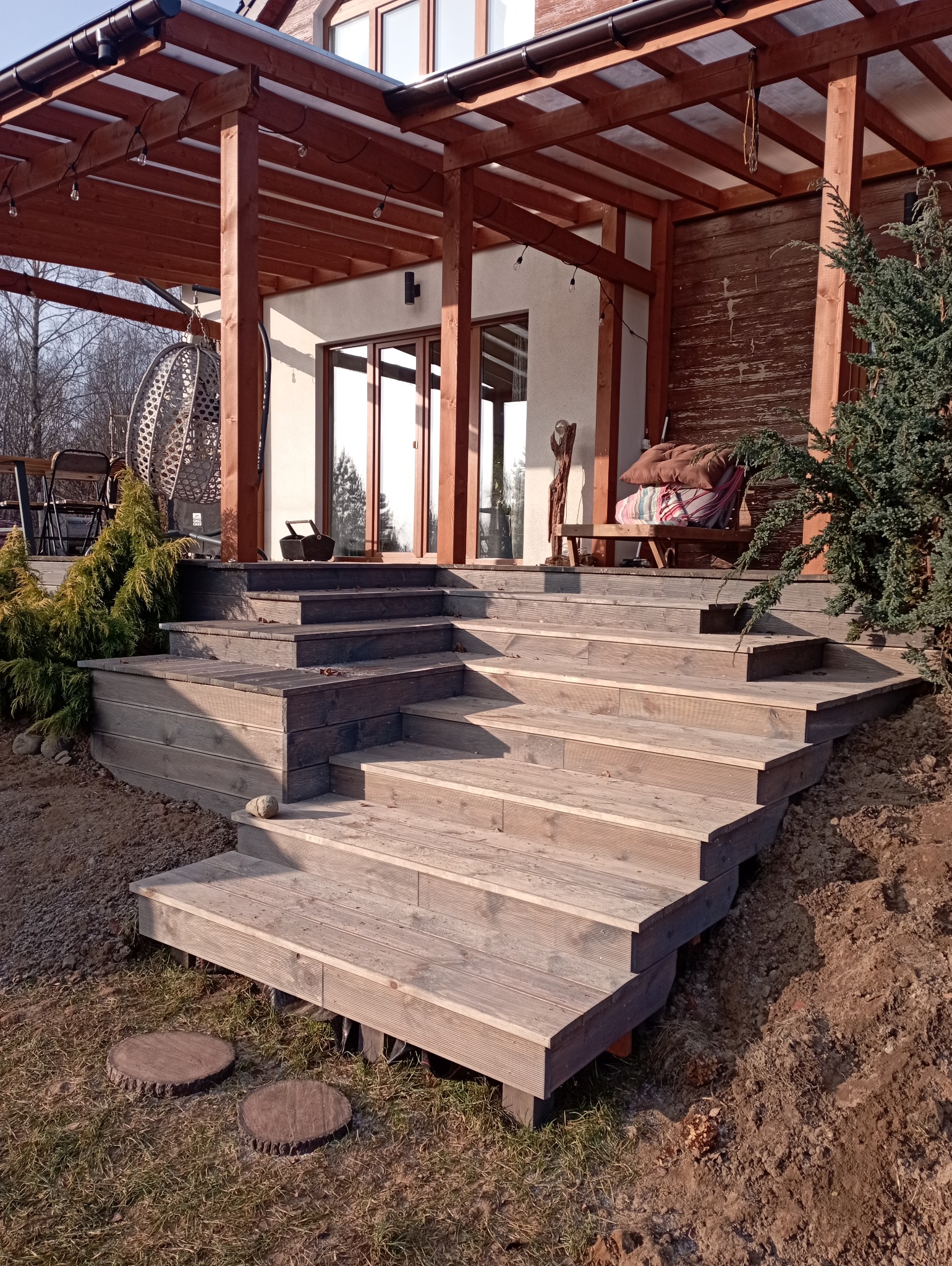 Drewniane schody tarasowe prowadzące do domu z pergolą, widoczne detale konstrukcyjne i naturalne otoczenie. Ujęcie z dołu podkreśla solidność wykonania.