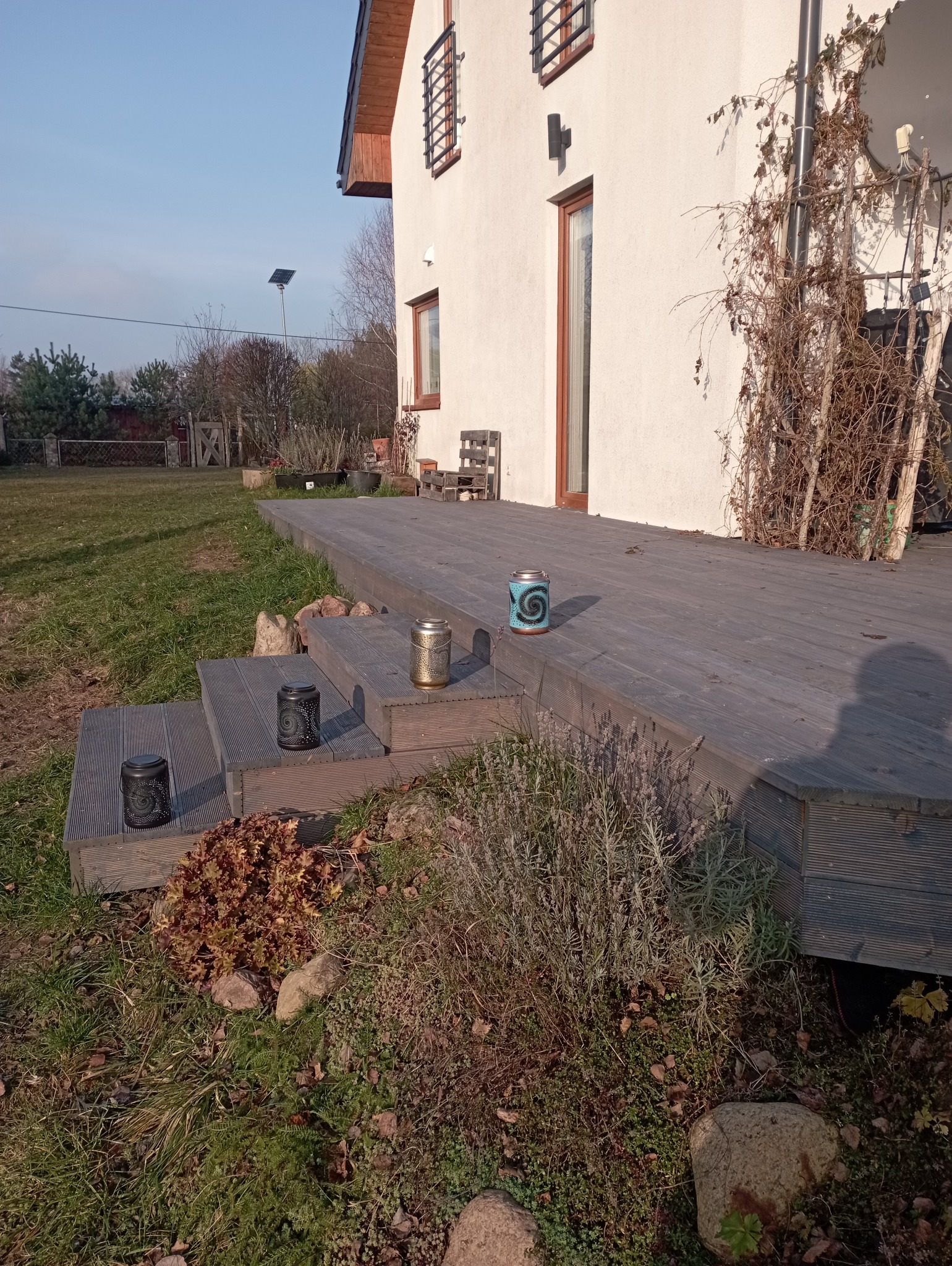 Drewniany taras z trzema stopniami, na których stoją dekoracyjne lampiony. W tle biały dom z drewnianymi oknami i balkonem porośniętym suchymi pnączami.