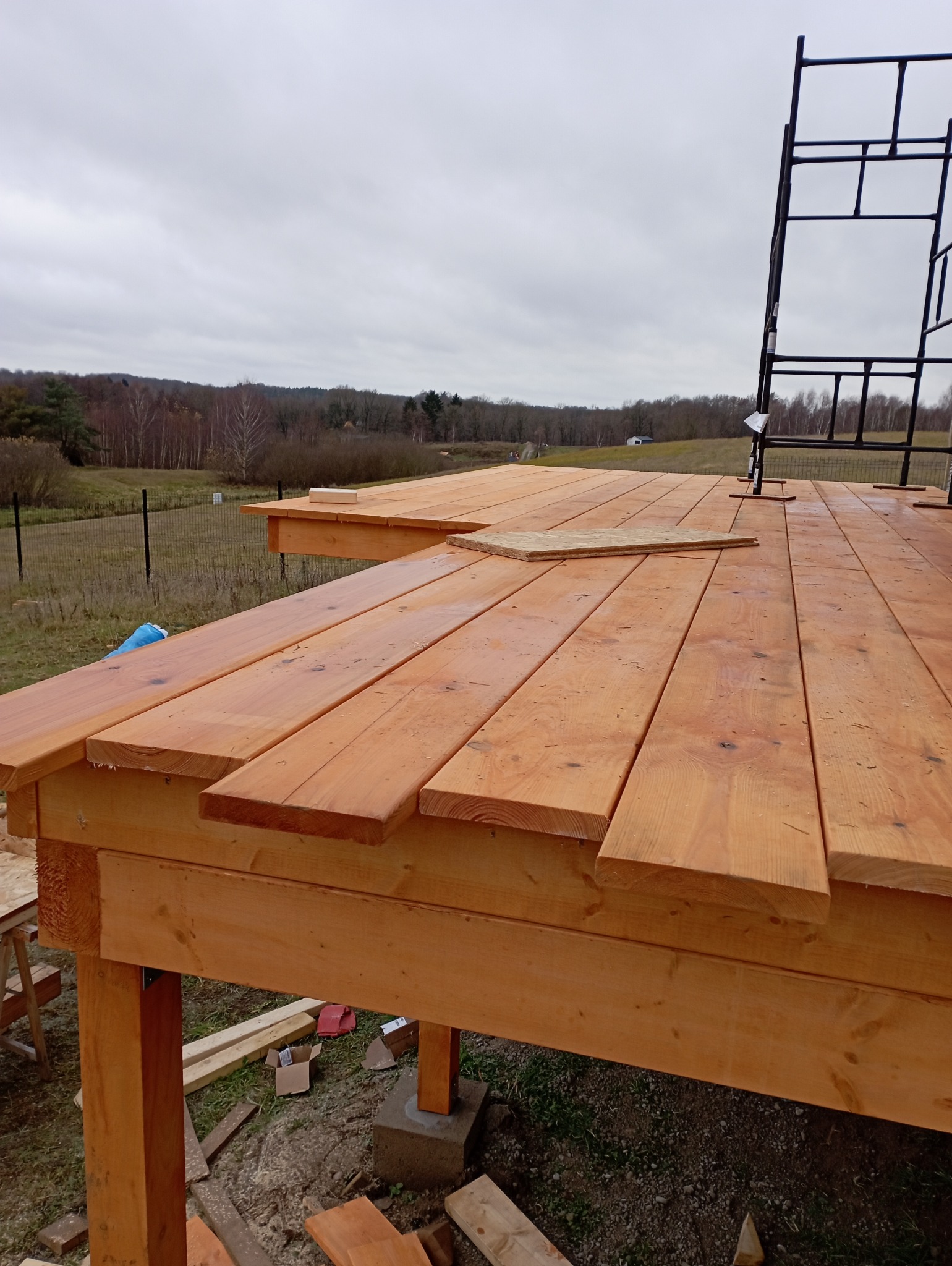Montaż drewnianego tarasu z desek o ciepłym odcieniu, widoczne elementy konstrukcyjne i rusztowanie w tle, na zewnątrz, pochmurny dzień. Częściowo ułożona podłoga.