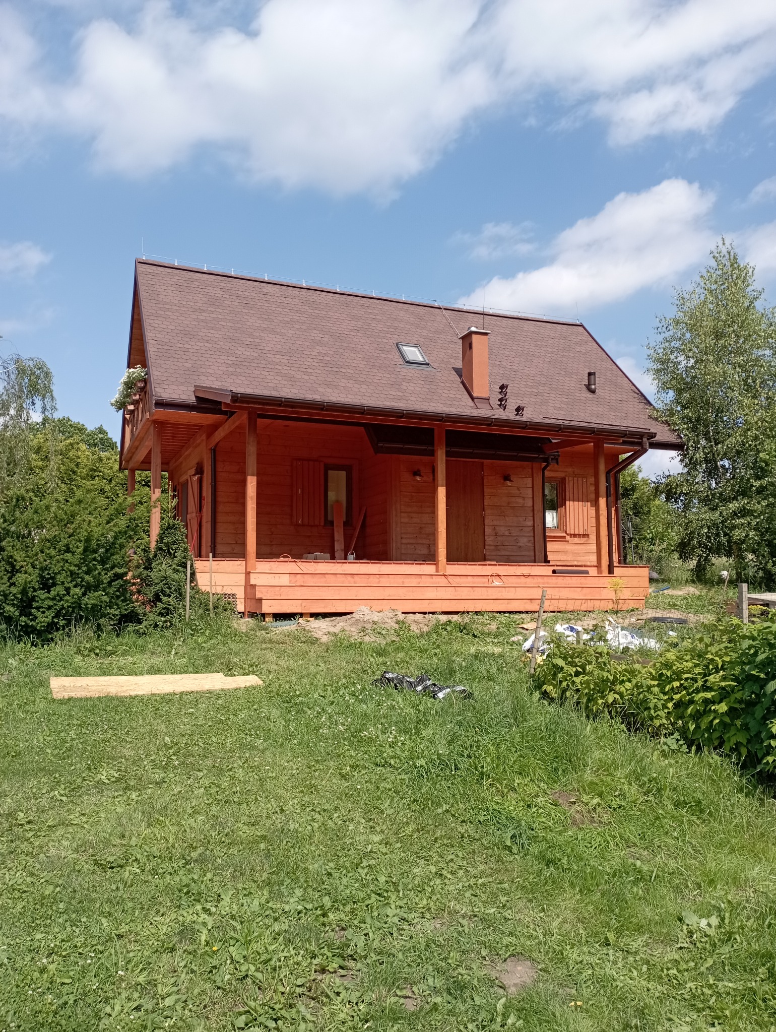 Drewniany dom szkieletowy z brązowym dachem, widoczny z zewnątrz, otoczony zielenią. Elementy wykończeniowe w kolorze drewna. Widoczne kwiaty w skrzynkach na balkonie.