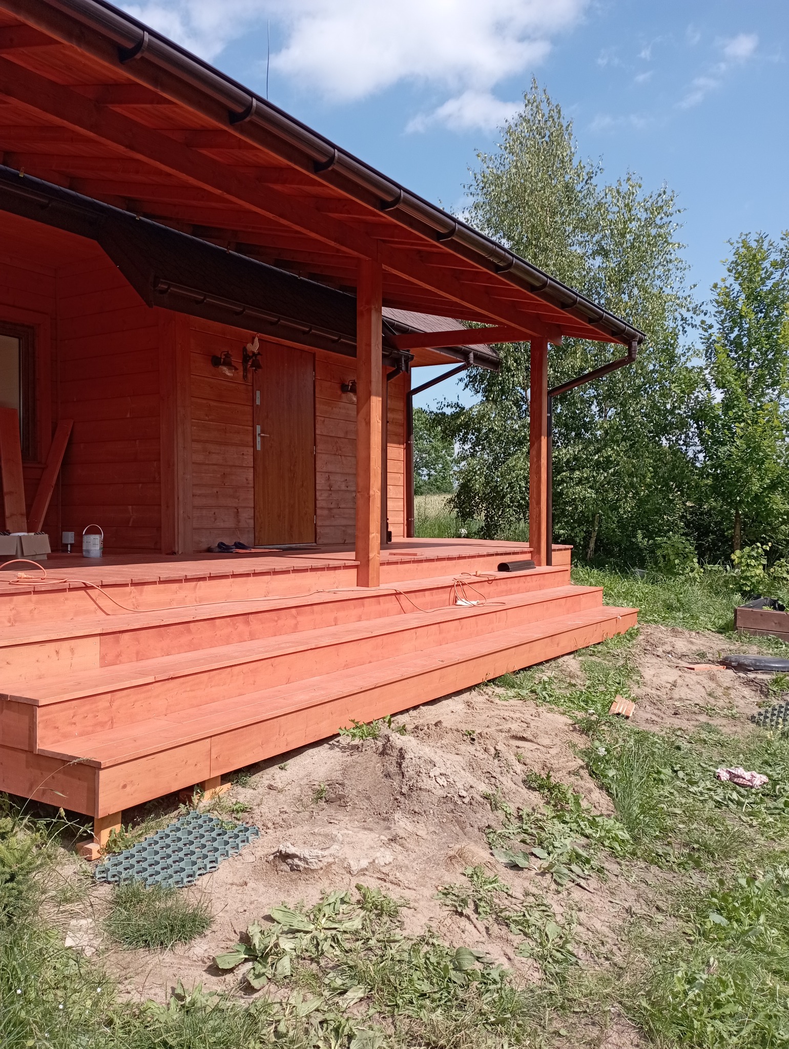 Nowy, drewniany taras z zadaszeniem przy domu, pomalowany na ciepły, pomarańczowy kolor. Widoczne schody wejściowe i elementy konstrukcyjne. W tle zieleń drzew i błękitne niebo.