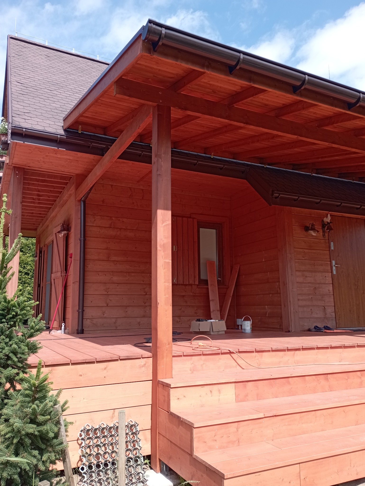 Ciepły odcień drewna domku z zadaszonym tarasem, widoczne schody wejściowe i elementy konstrukcyjne. Częściowo widoczne narzędzia i materiały budowlane.