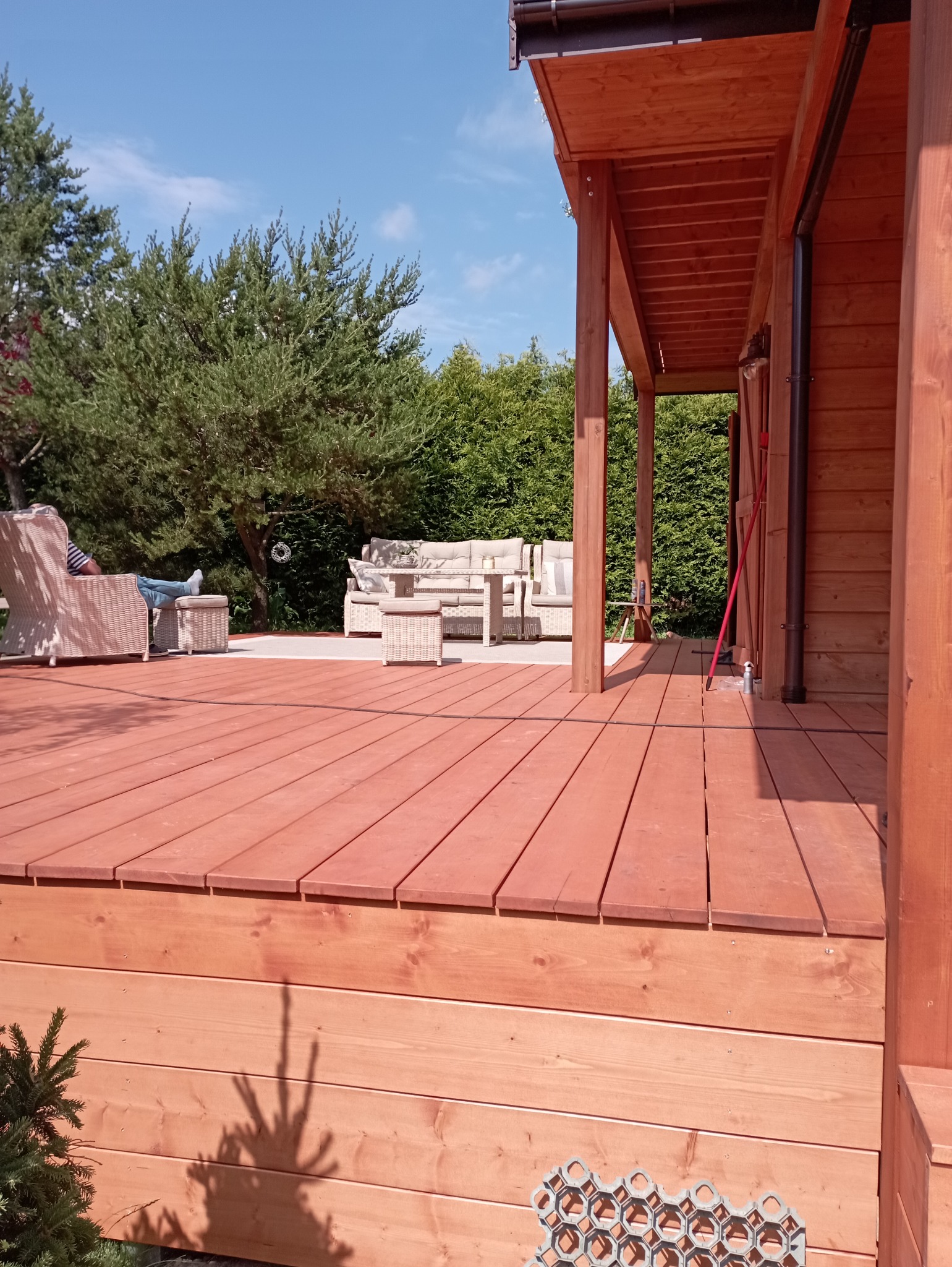 Ciepły odcień drewna na tarasie z meblami ogrodowymi i relaksującą osobą w tle, otoczony zielenią. Widoczne elementy konstrukcyjne z drewna.