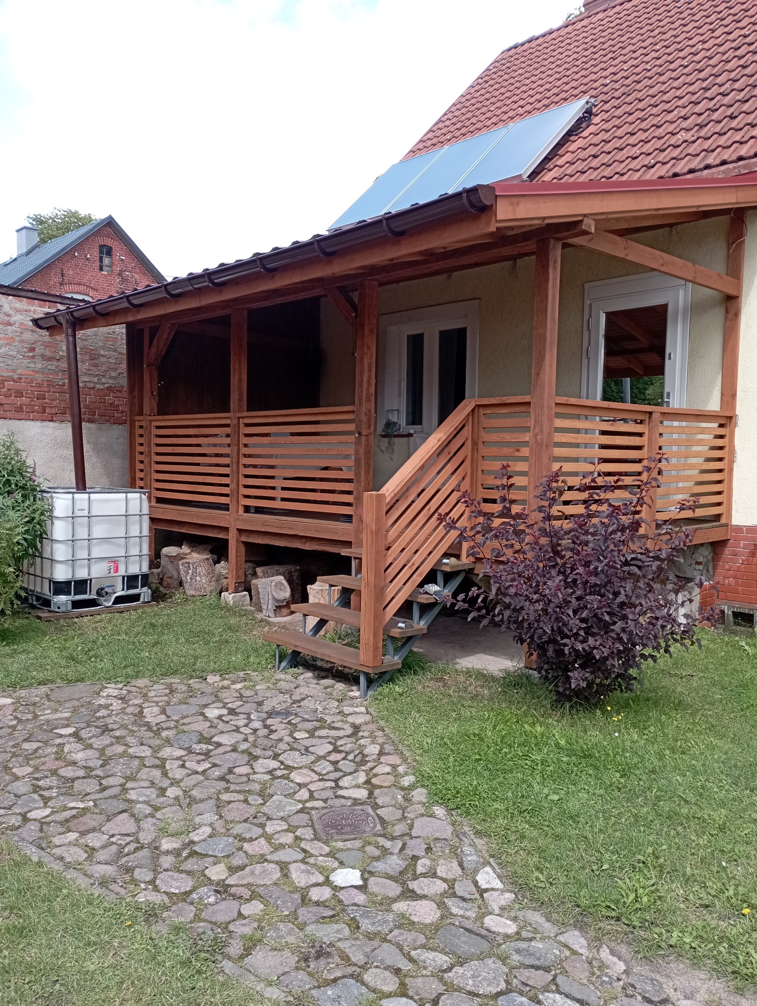 Zadaszony taras z drewnianą balustradą i schodami, widok z zewnątrz. Obok zbiornik na wodę i ścieżka z kamienia polnego. Na dachu panele słoneczne.