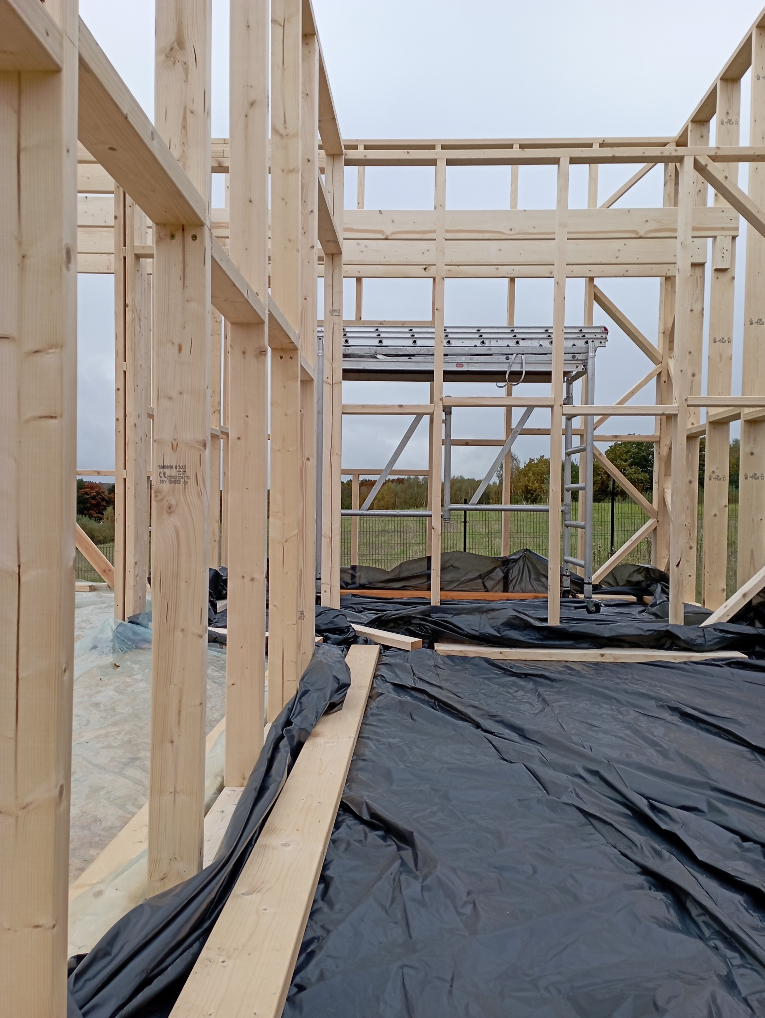 Surowy szkielet domu w budowie, z widocznymi drewnianymi belkami i rusztowaniem w tle. Czarna folia ochronna na podłożu. Widok z bliska na konstrukcję.