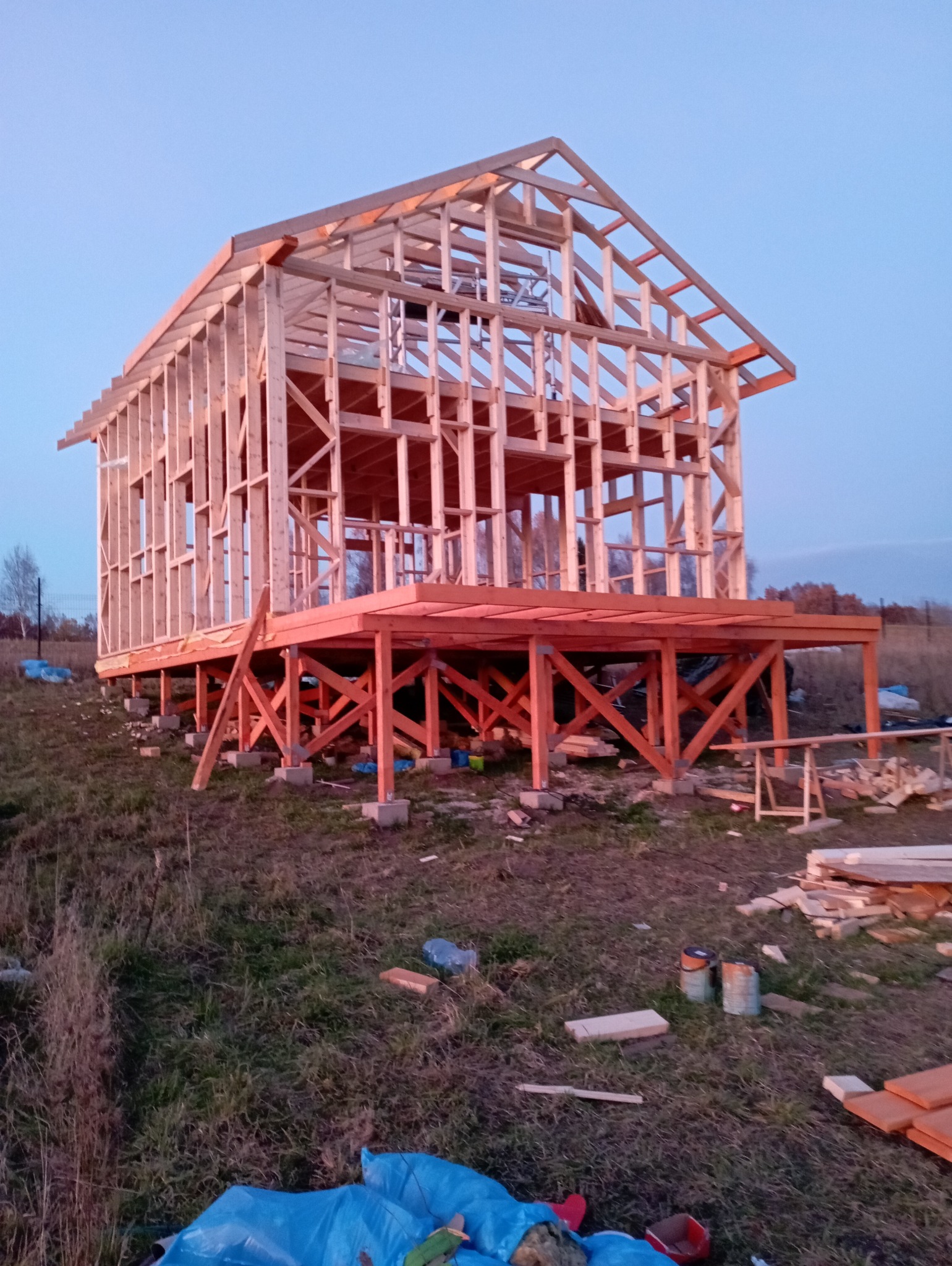 Szkielet drewniany domu w trakcie budowy, widoczny na tle nieba o zmierzchu. Konstrukcja osadzona na betonowych fundamentach, z widocznymi elementami drewnianymi.